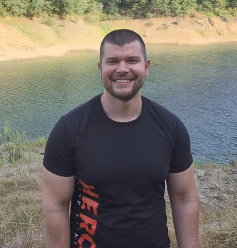 Smiling man in a black shirt standing outdoors near a body of water with trees in the background.