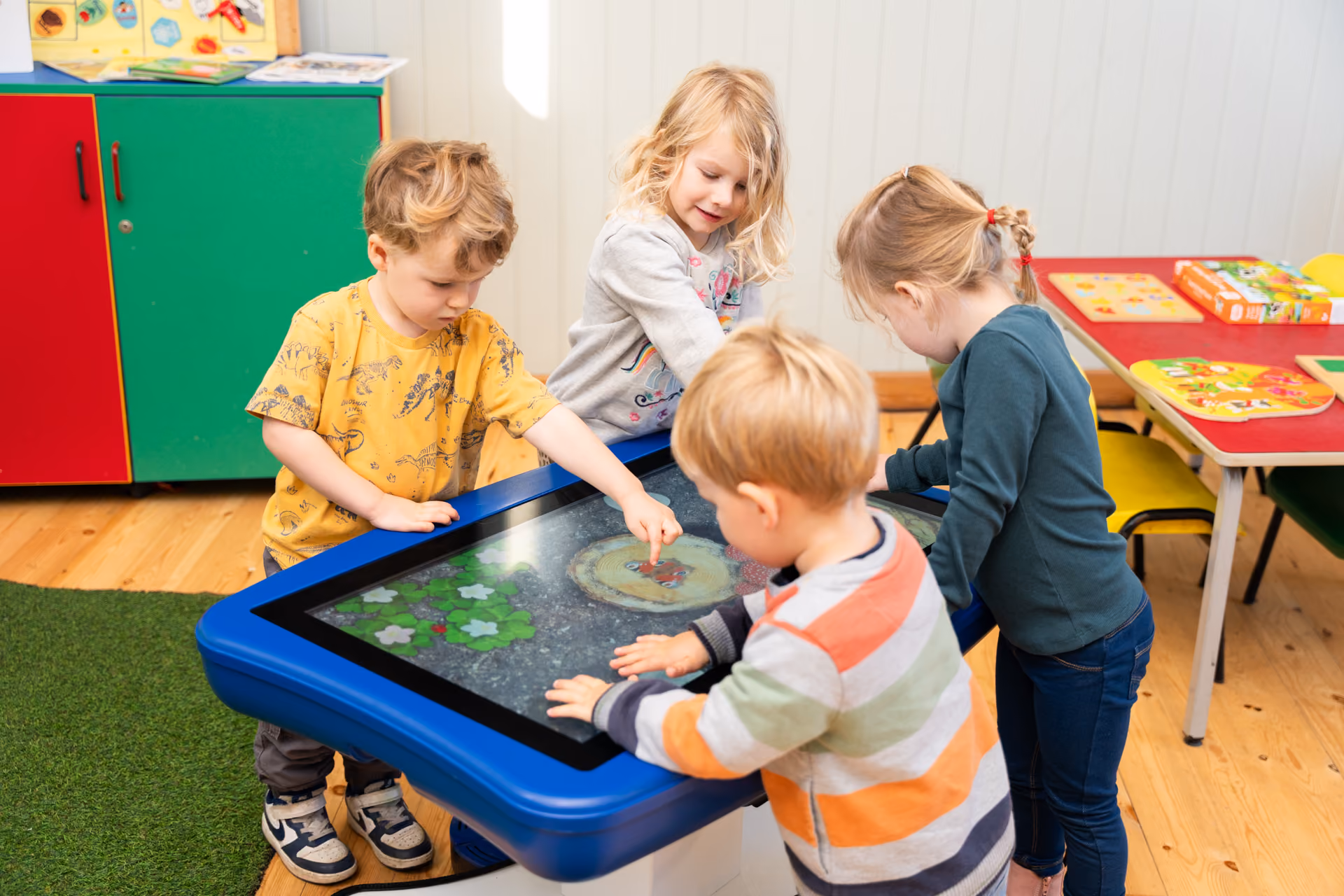 Four young children playing together on a large blue interactive touchscreen table in a classroom.