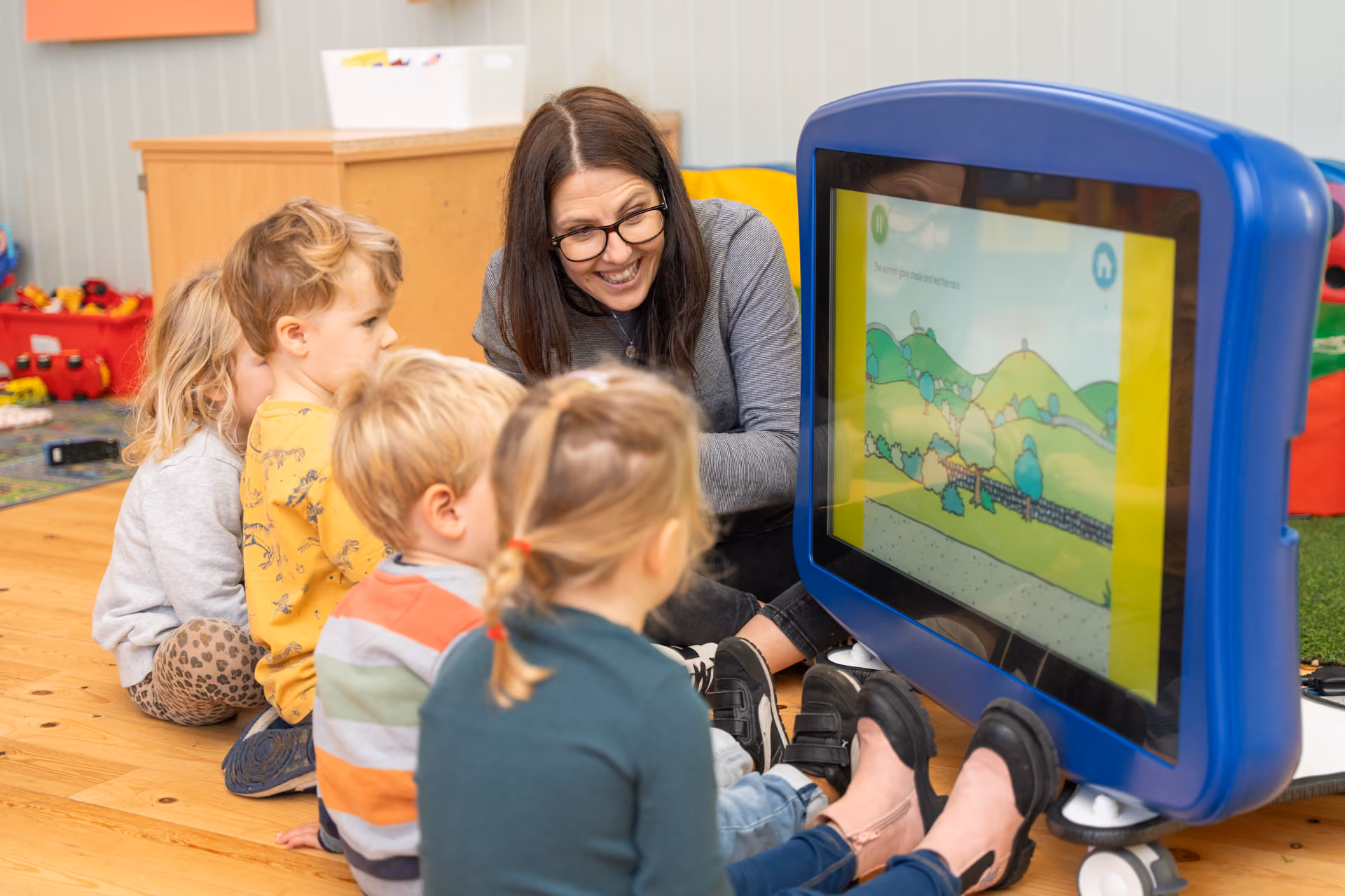 Teacher sitting on the floor with a group of young children watching a large interactive screen displaying a colorful hills and trees scene.