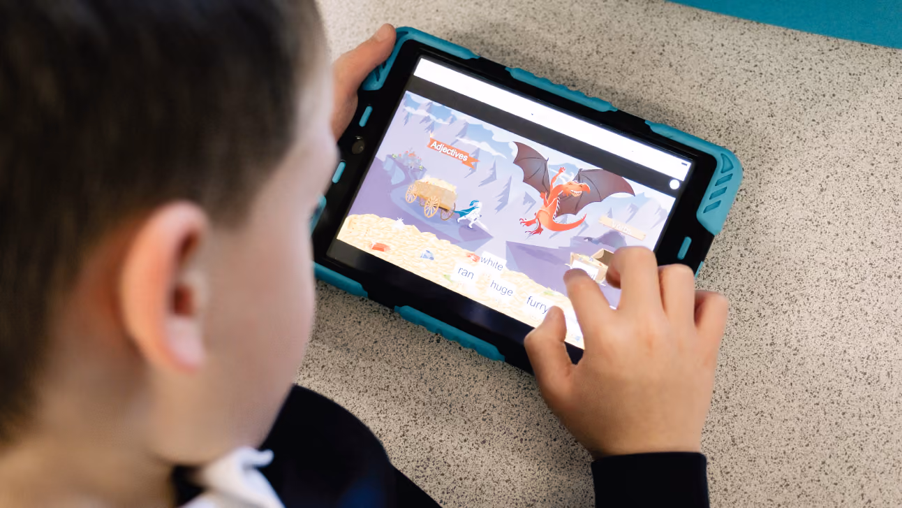 Child interacting with a tablet displaying an educational game about adjectives and verbs featuring a dragon and treasure.