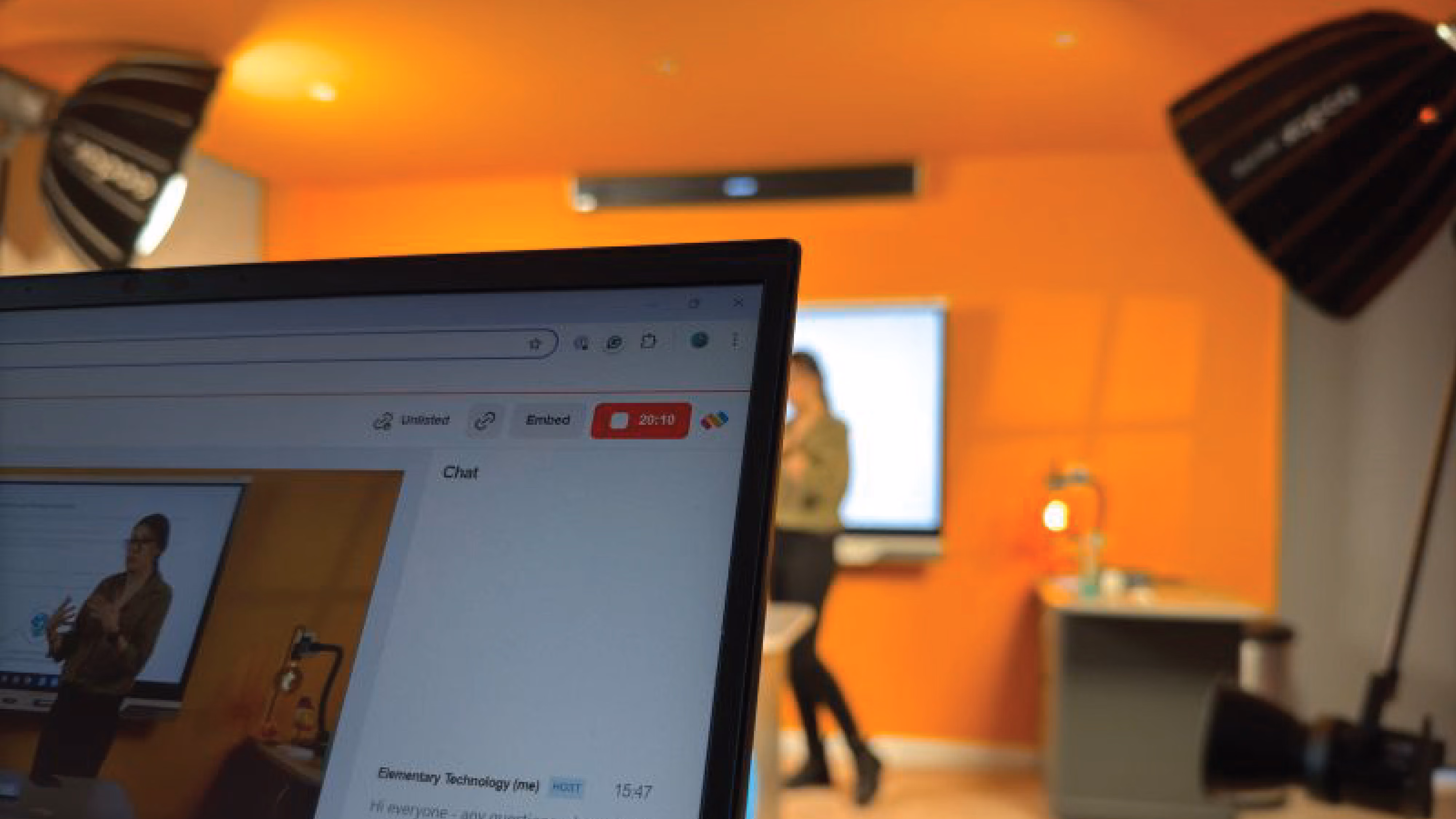 Close-up of a laptop screen showing a live video of a woman presenting in a room with an orange wall and studio lights.