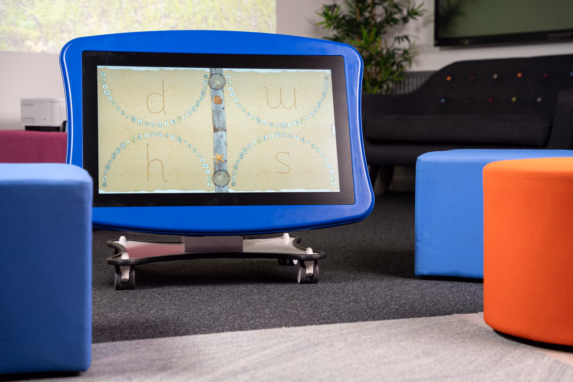 Interactive touchscreen device with a blue frame displaying letters d, h, w, and s surrounded by alphabet letters on a sandy background, in a room with colorful cubes and a dark sofa.