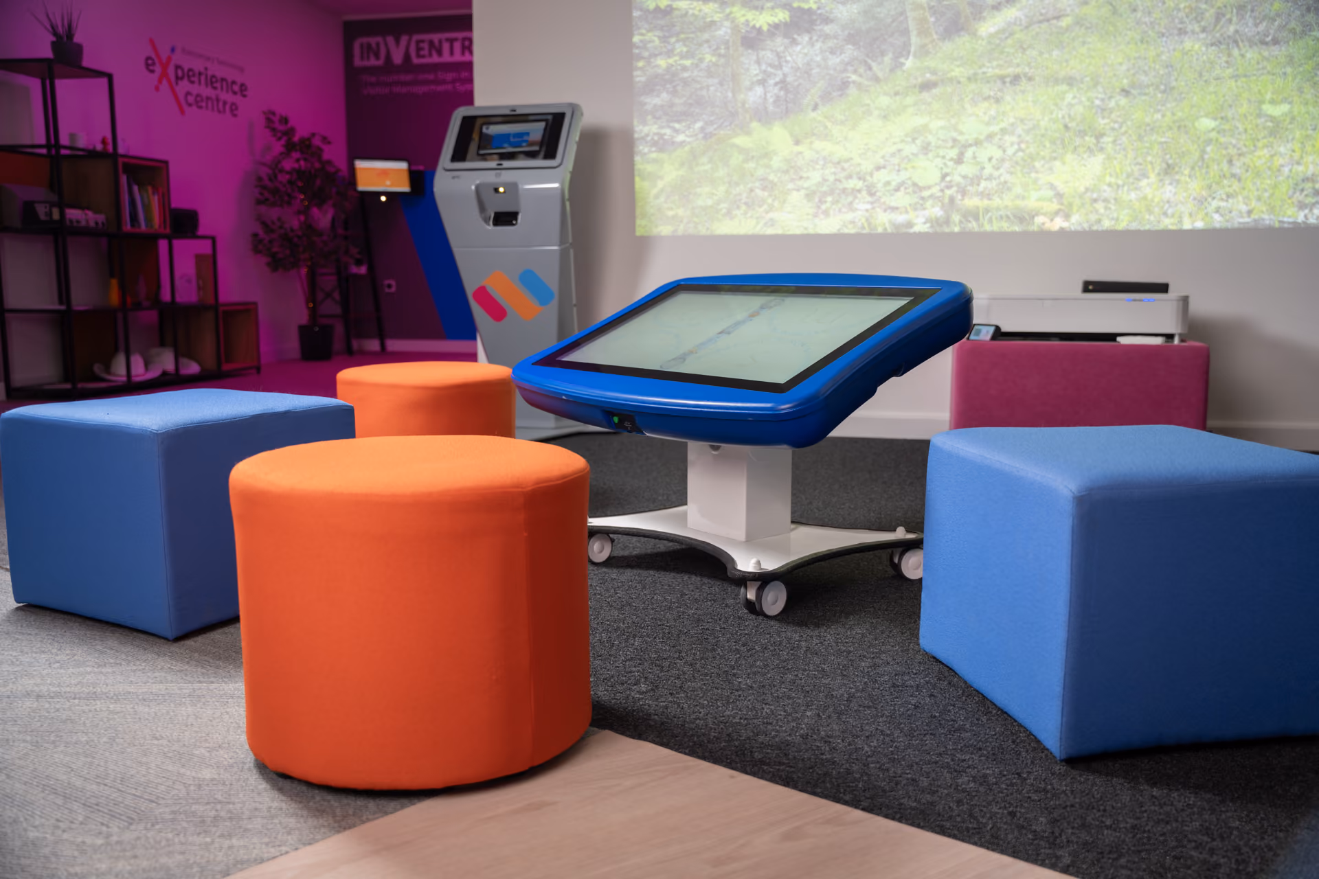 Interactive blue digital touchscreen table surrounded by colorful orange and blue soft seating cubes in a modern experience center room.
