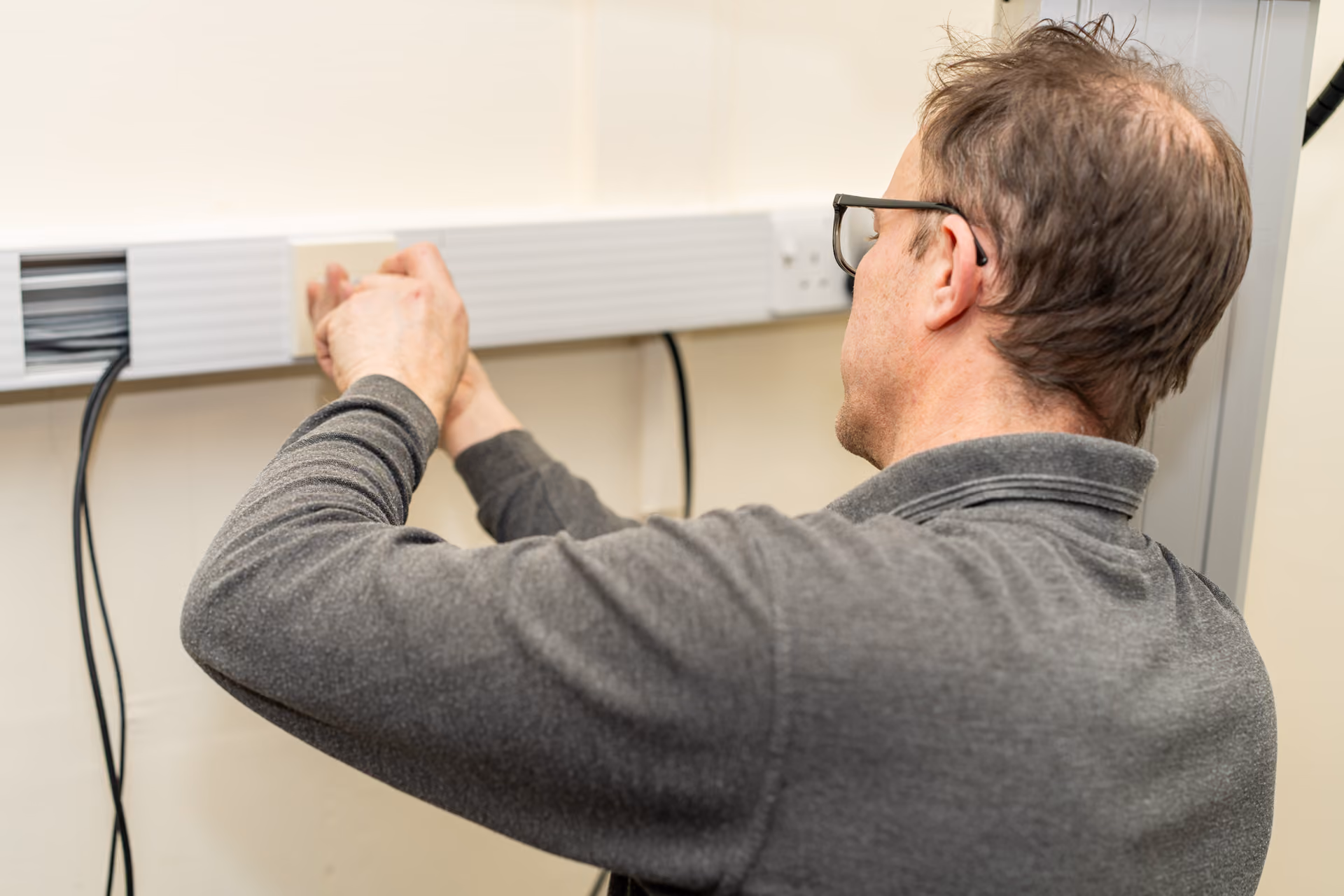 Man wearing glasses and a gray sweater working on cable management inside a plastic wall-mounted conduit.