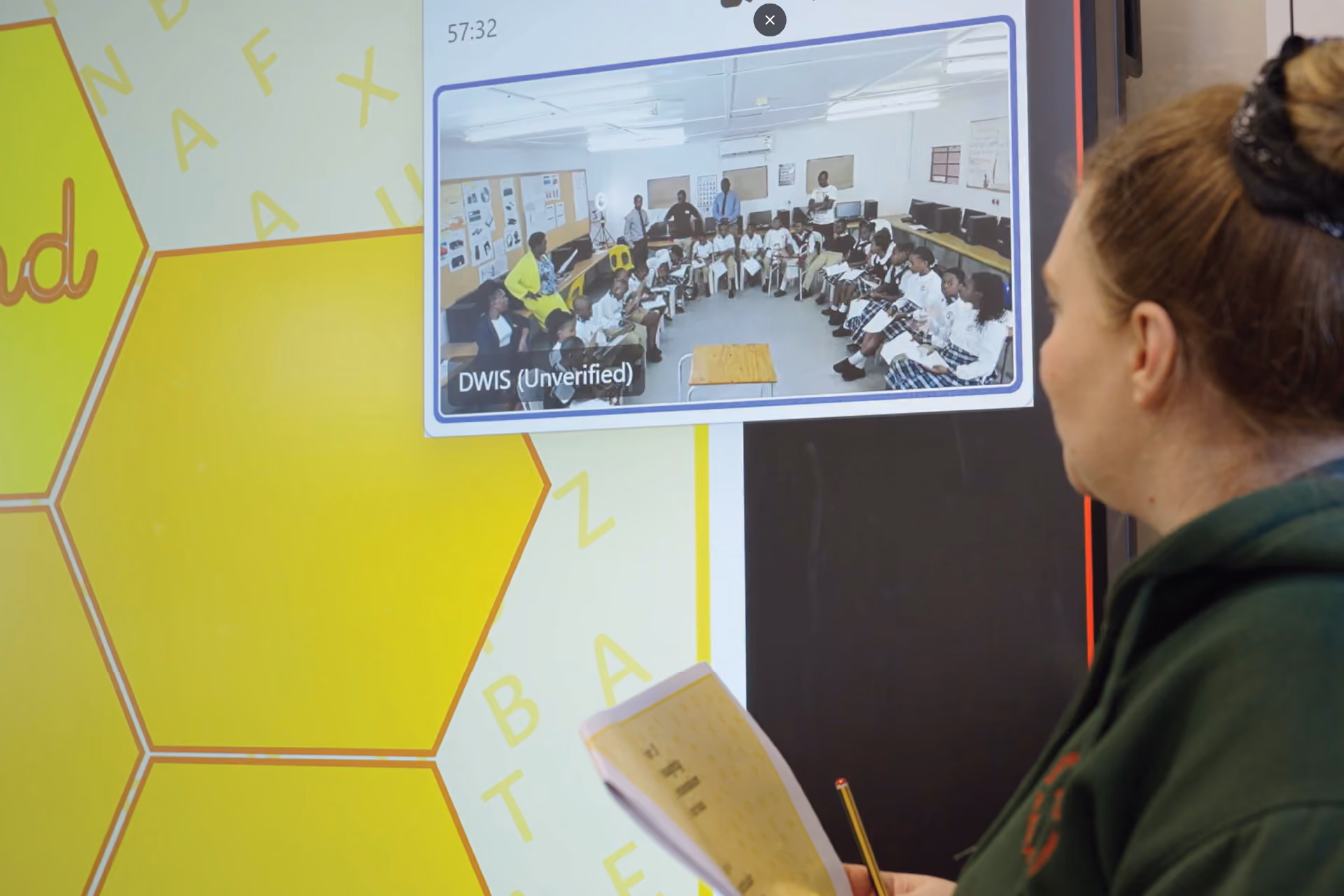 Woman holding a pencil and paper, looking at a screen showing a classroom of students seated in a circle with teachers standing around.