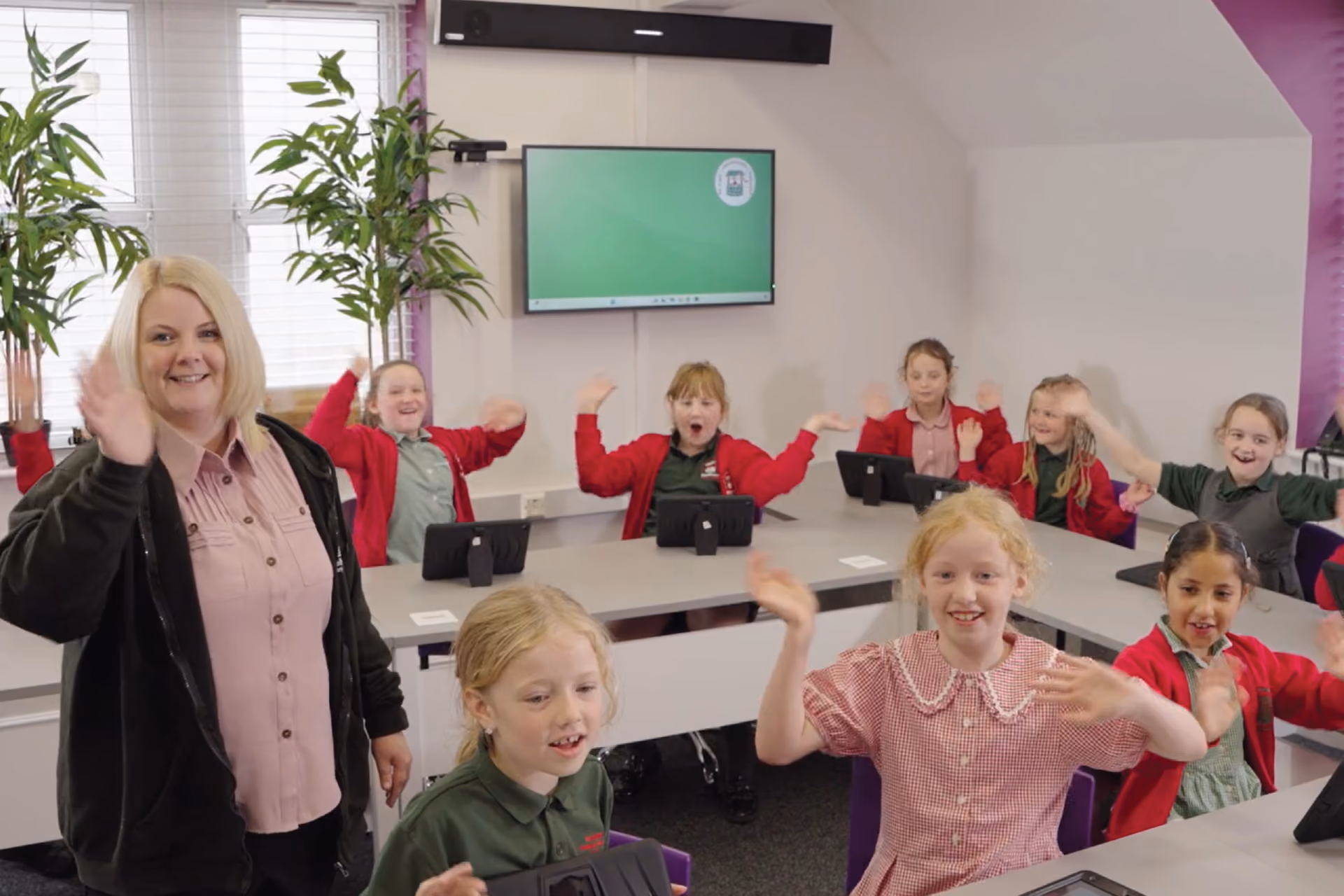 Teacher and elementary school children in a classroom waving and smiling, with tablets on the desks and a screen on the wall.