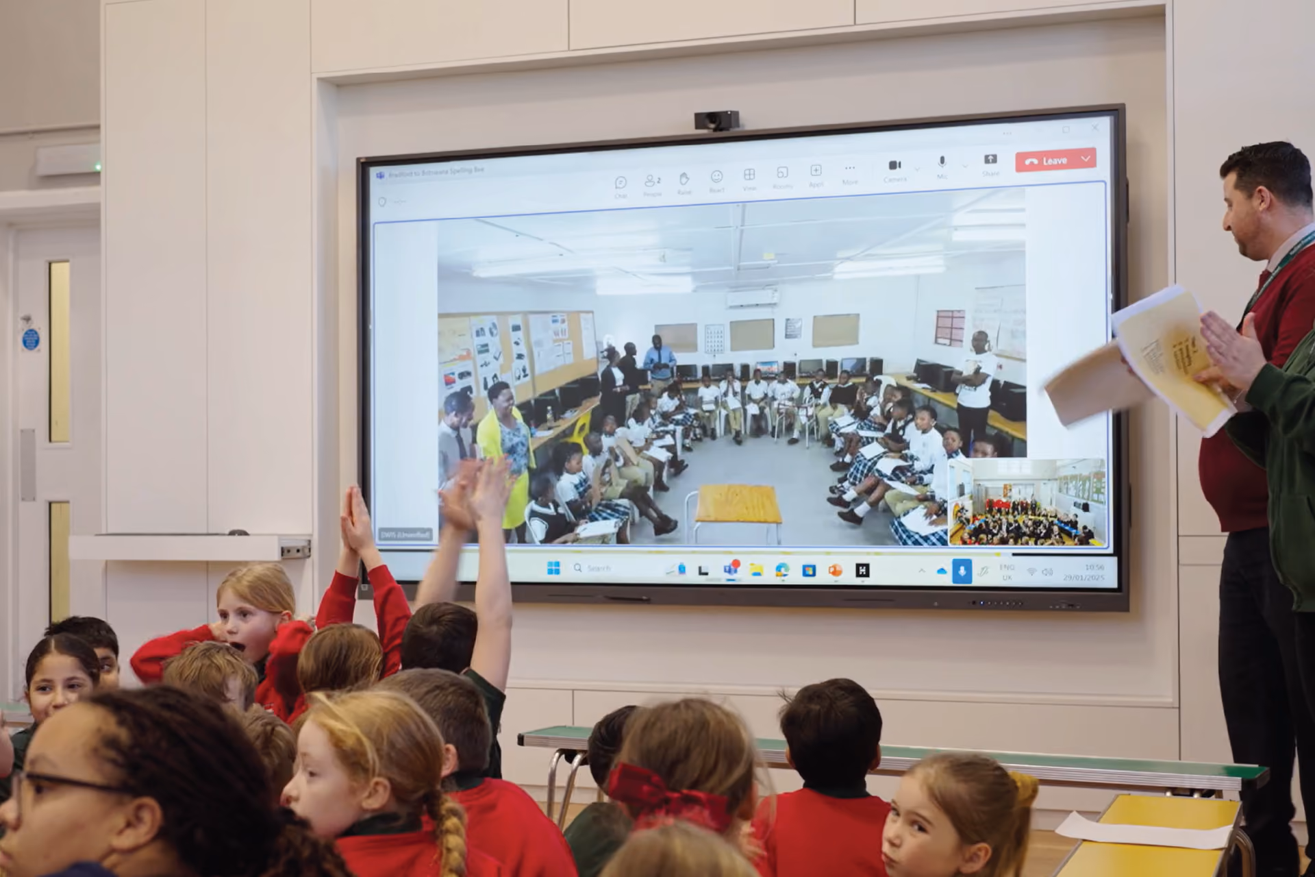 Children in a classroom watching a large screen showing another classroom with students and teachers participating virtually.