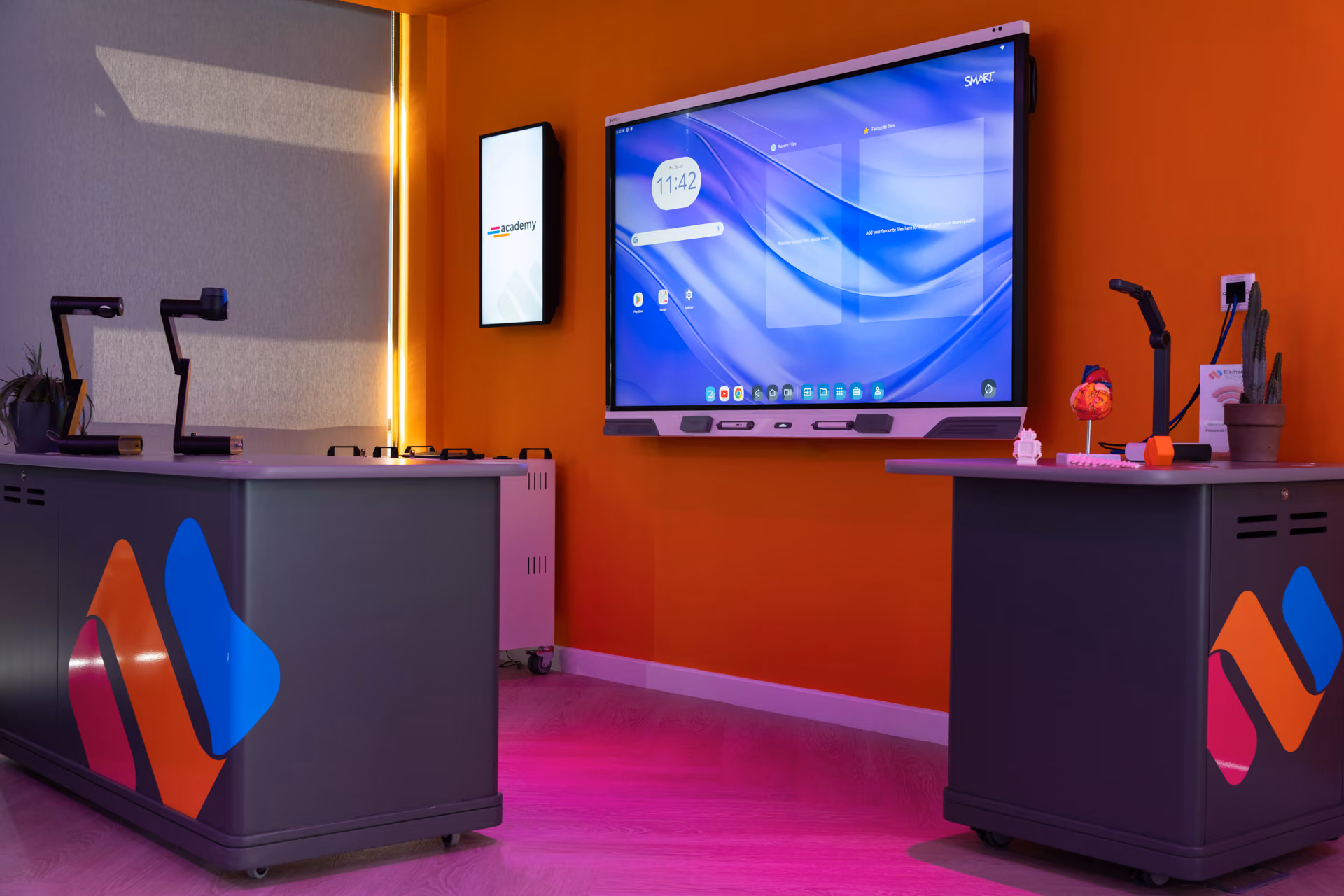 Modern classroom with two gray mobile desks featuring colorful logos, a large interactive SMART board on an orange wall, and desk-mounted document cameras.