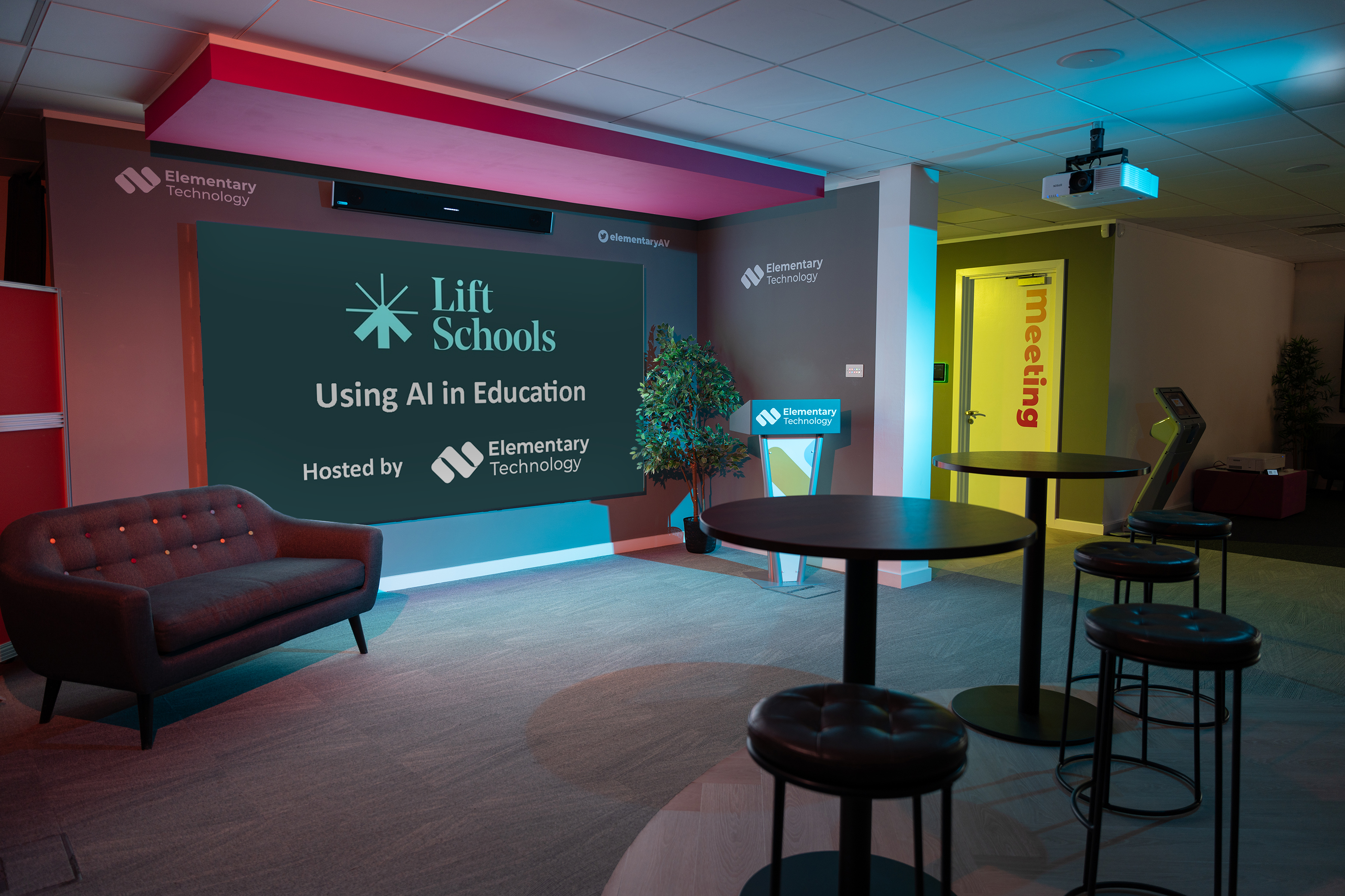 Modern meeting room with a screen showing 'Lift Schools Using AI in Education Hosted by Elementary Technology' and several stools and tables.