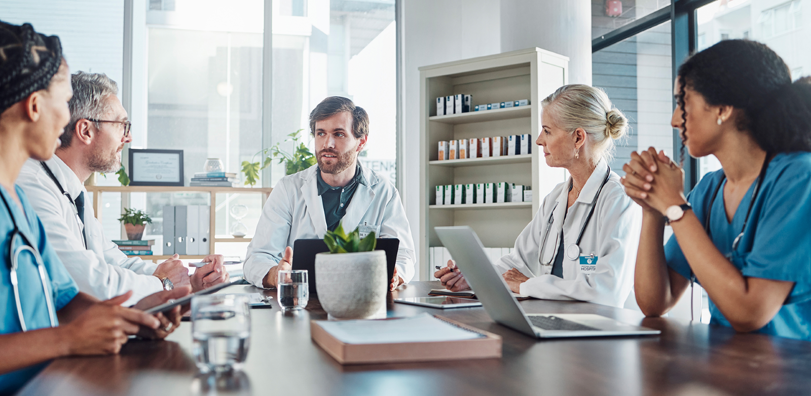 A group of doctors in a corner office lit up by natural light with their laptops open having a meeting.