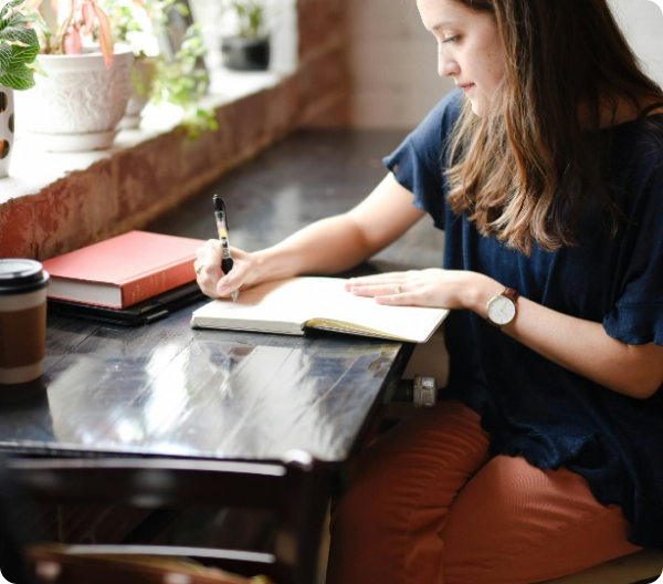 Mulher sentada em mesa junto à janela, escrevendo em um caderno aberto com caneta, com livro vermelho e copo de café na mesa.