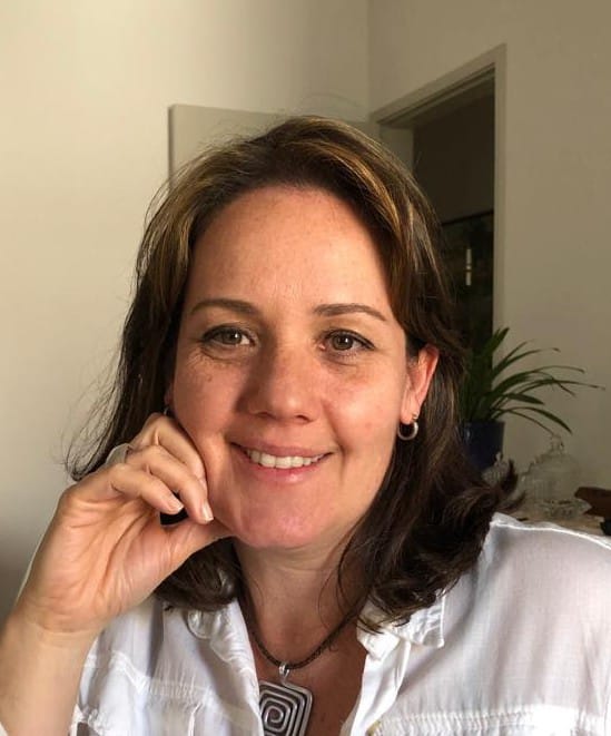 Mulher sorridente com cabelo castanho e luzes loiras, usando blusa branca e colar, posando com a mão apoiada no rosto.