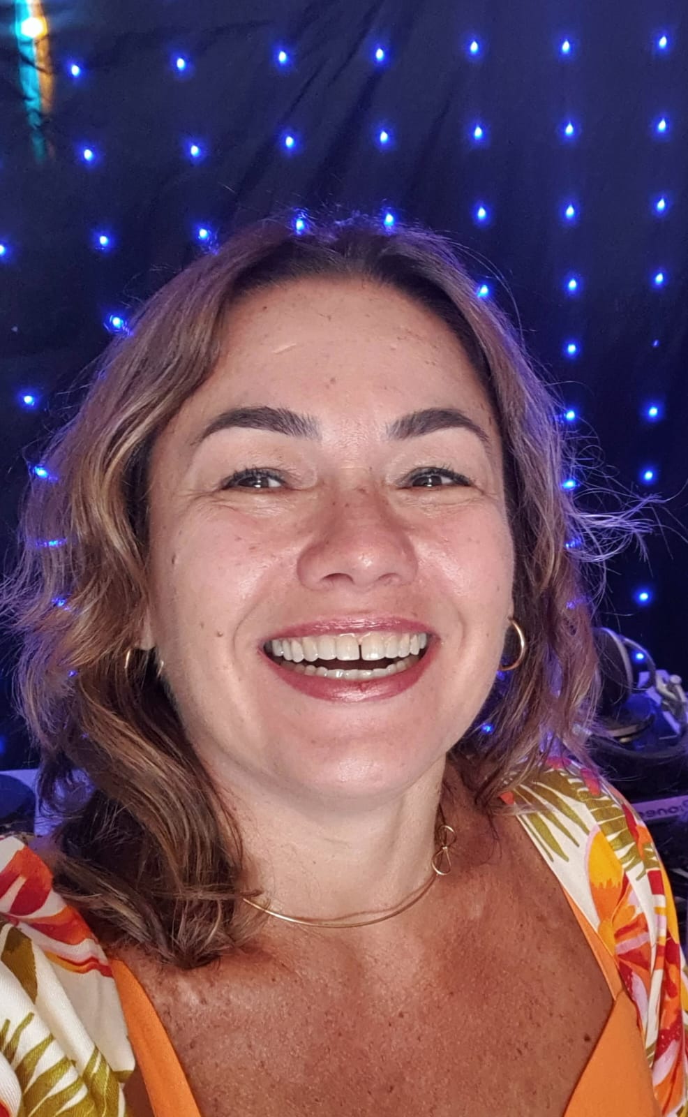 Mulher sorridente com cabelo ondulado castanho-claro usando brincos de argola dourados e blusa laranja com estampa floral.