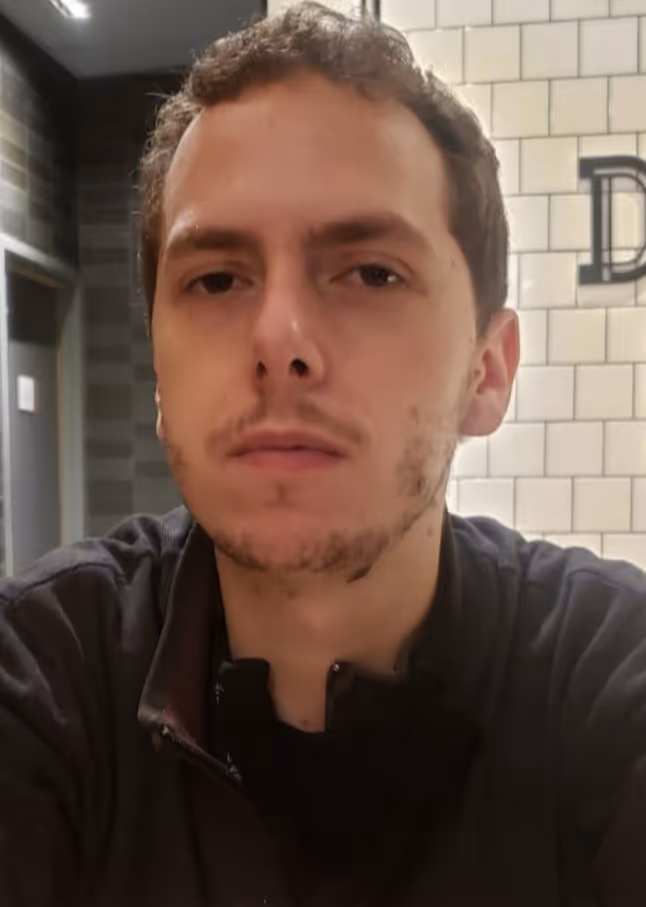 Retrato de um homem jovem com expressão séria, cabelo curto e jaqueta preta, com parede de azulejos brancos ao fundo.