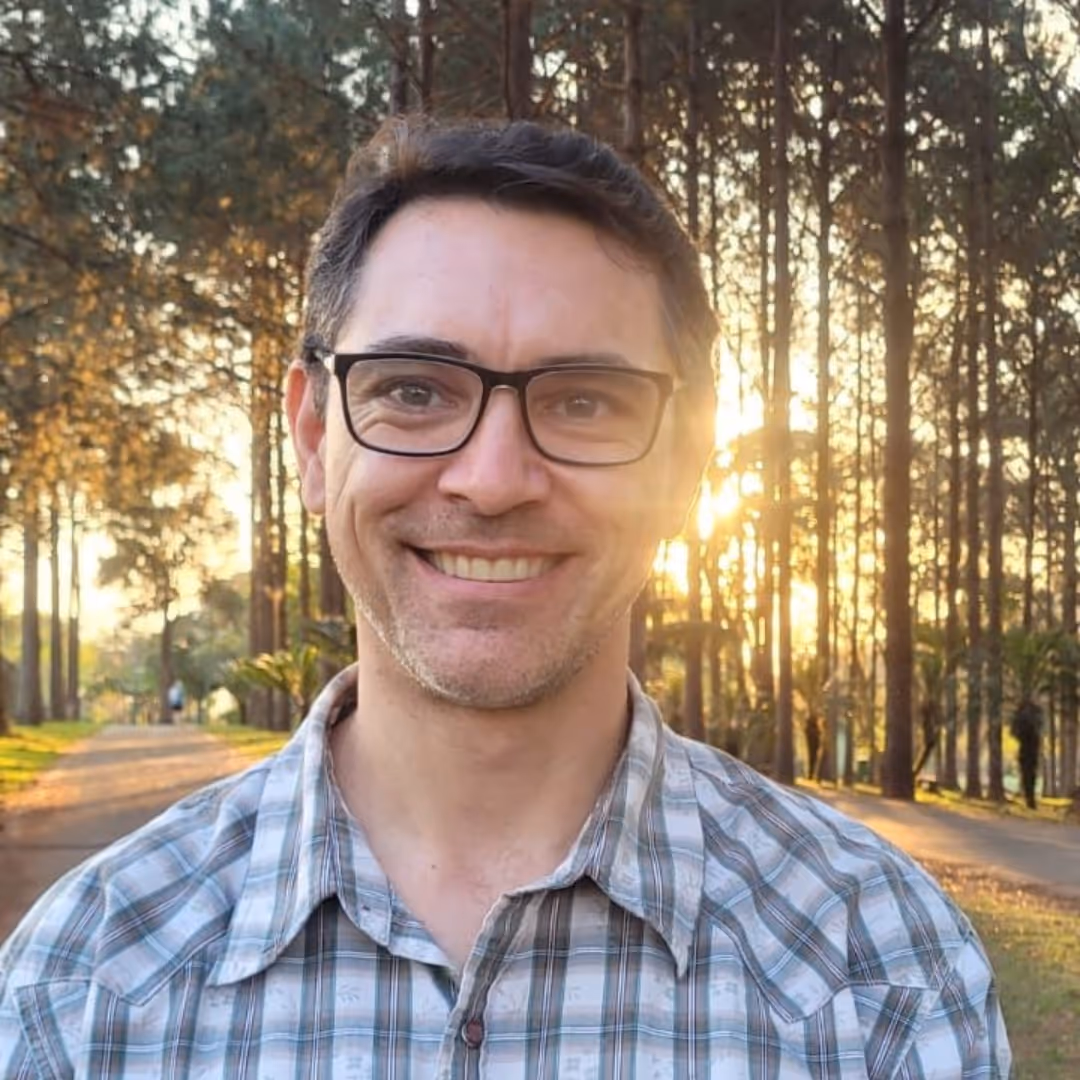 Homem sorridente de óculos com camisa xadrez em parque ao pôr do sol entre árvores.