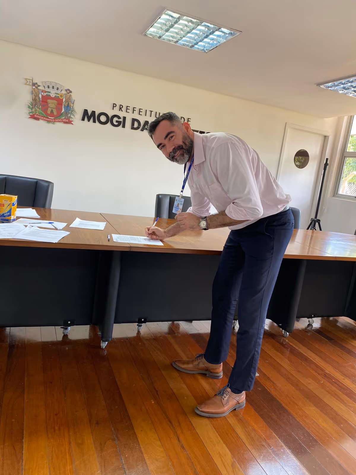 Homem de camisa rosa clara, calça escura e sapatos marrons assinando um documento em uma mesa de madeira em sala da Prefeitura de Mogi das Cruzes.