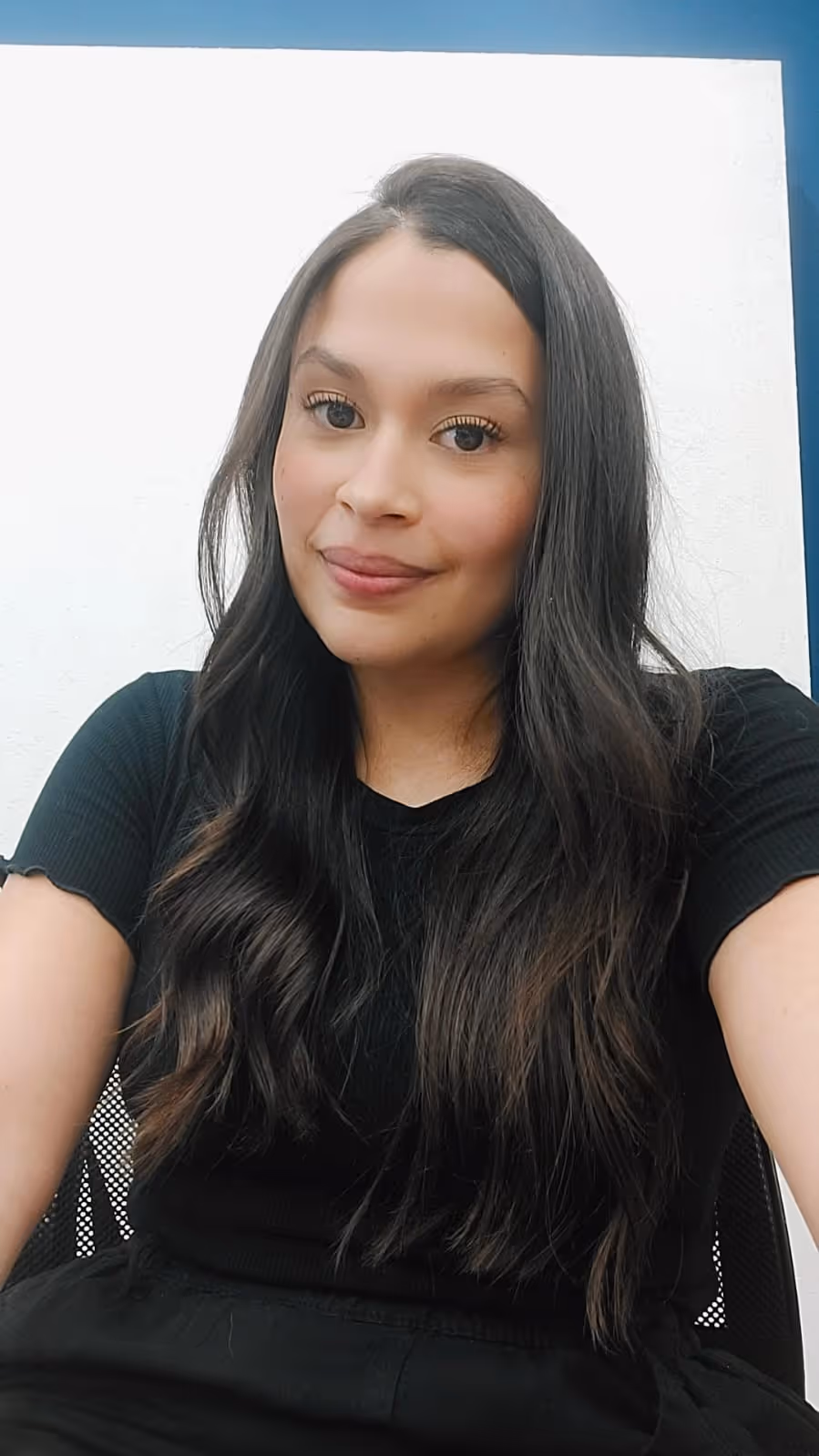 Mulher sorrindo com cabelos longos e ondulados, usando camiseta preta, sentada em cadeira com fundo branco.