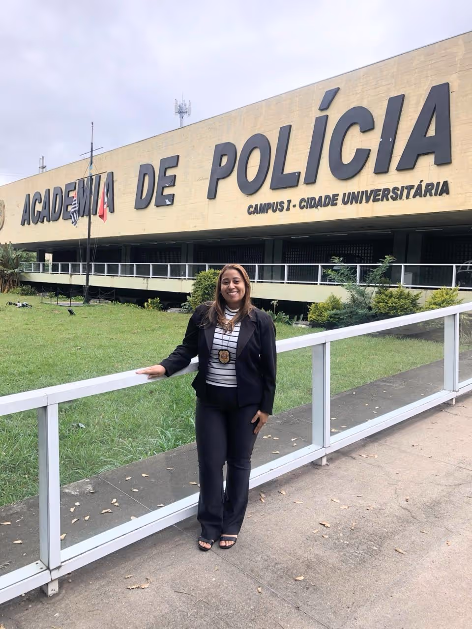 Mulher sorridente com crachá da polícia, em pé ao lado de uma grade branca em frente ao prédio da Academia de Polícia, Campus 1, Cidade Universitária.