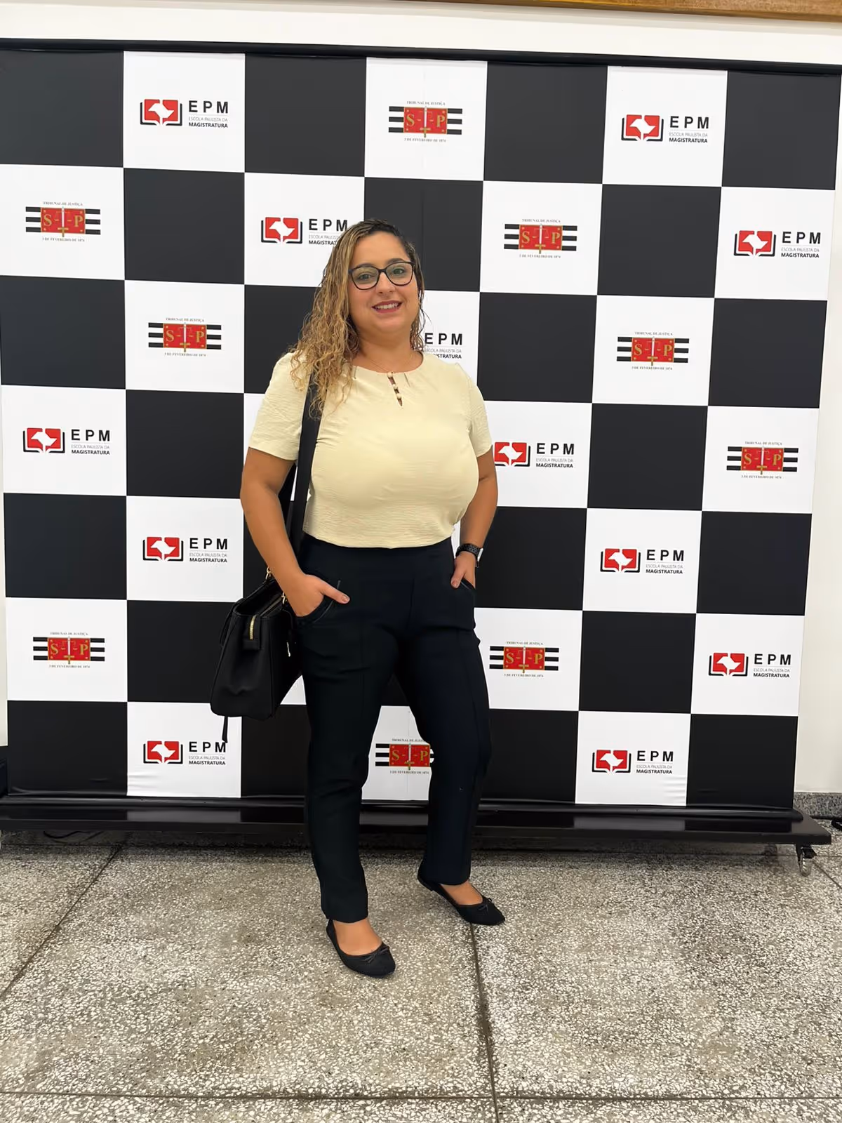 Mulher sorridente com óculos, camiseta branca, calça preta e bolsa preta, posando em frente a um painel quadriculado com logos da EPM e Tribunal de Justiça de São Paulo.