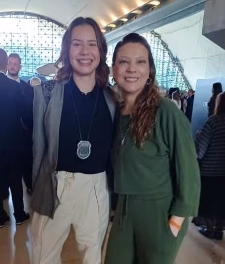 Duas mulheres sorrindo abraçadas em um ambiente interno iluminado, uma usando crachá pendurado no pescoço.