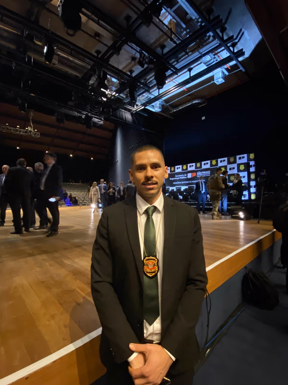 Homem vestido formalmente com terno preto, gravata verde e distintivo pendurado no pescoço, posando em um palco com outras pessoas ao fundo em um evento oficial.