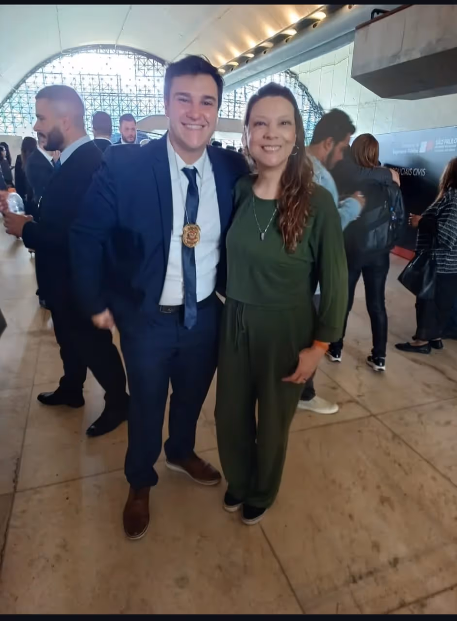 Homem de terno azul com distintivo pendurado no pescoço ao lado de mulher com macacão verde em ambiente interno com outras pessoas ao fundo.