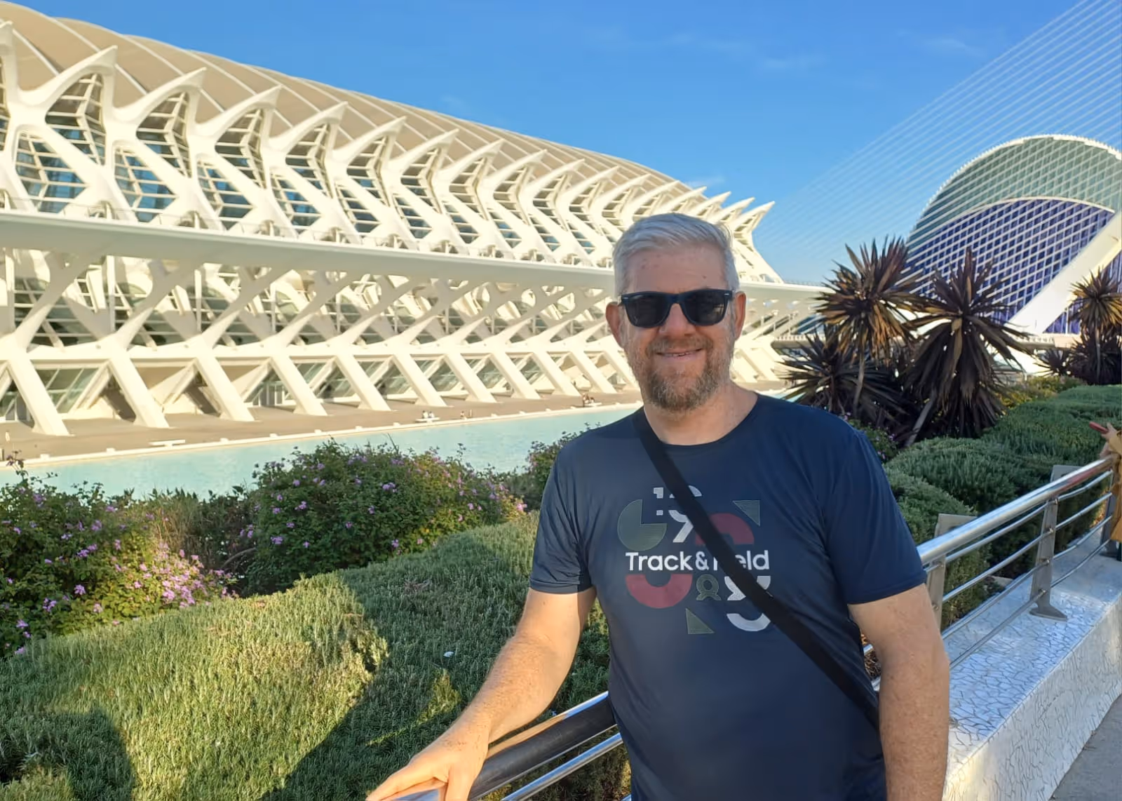 Homem de camiseta azul escura e óculos escuros encostado em um corrimão com fundo de arquitetura moderna e plantas.