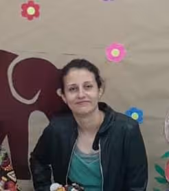 Mulher de cabelo preso usando jaqueta preta, posando sorridente em frente a um mural com desenhos de flores coloridas e parte de um elefante marrom.