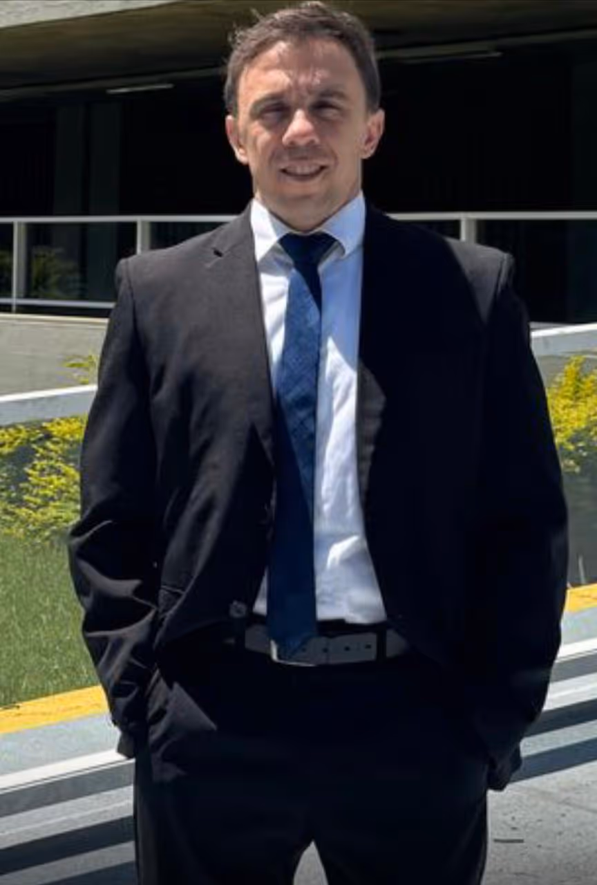 Homem vestindo terno preto, camisa branca e gravata azul, posando ao ar livre com mãos nos bolsos.