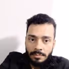 Retrato de um homem jovem com barba e cabelo curto encaracolado, usando camiseta preta, com fundo branco.