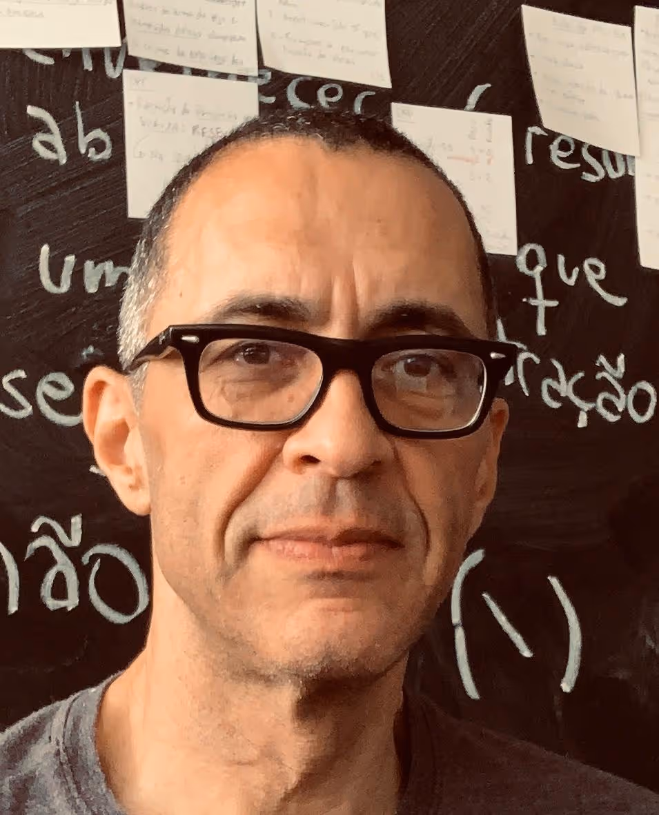 Homem de óculos com cabelos curtos e grisalhos, com quadro negro e anotações ao fundo.