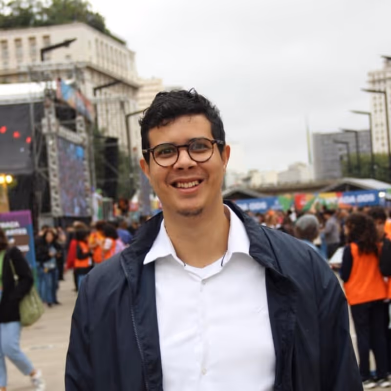 Homem sorridente com óculos e jaqueta escura em ambiente urbano com pessoas ao fundo.