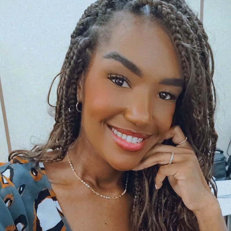 Mulher sorridente com tranças, usando colar delicado e anéis, apoiando o rosto na mão.