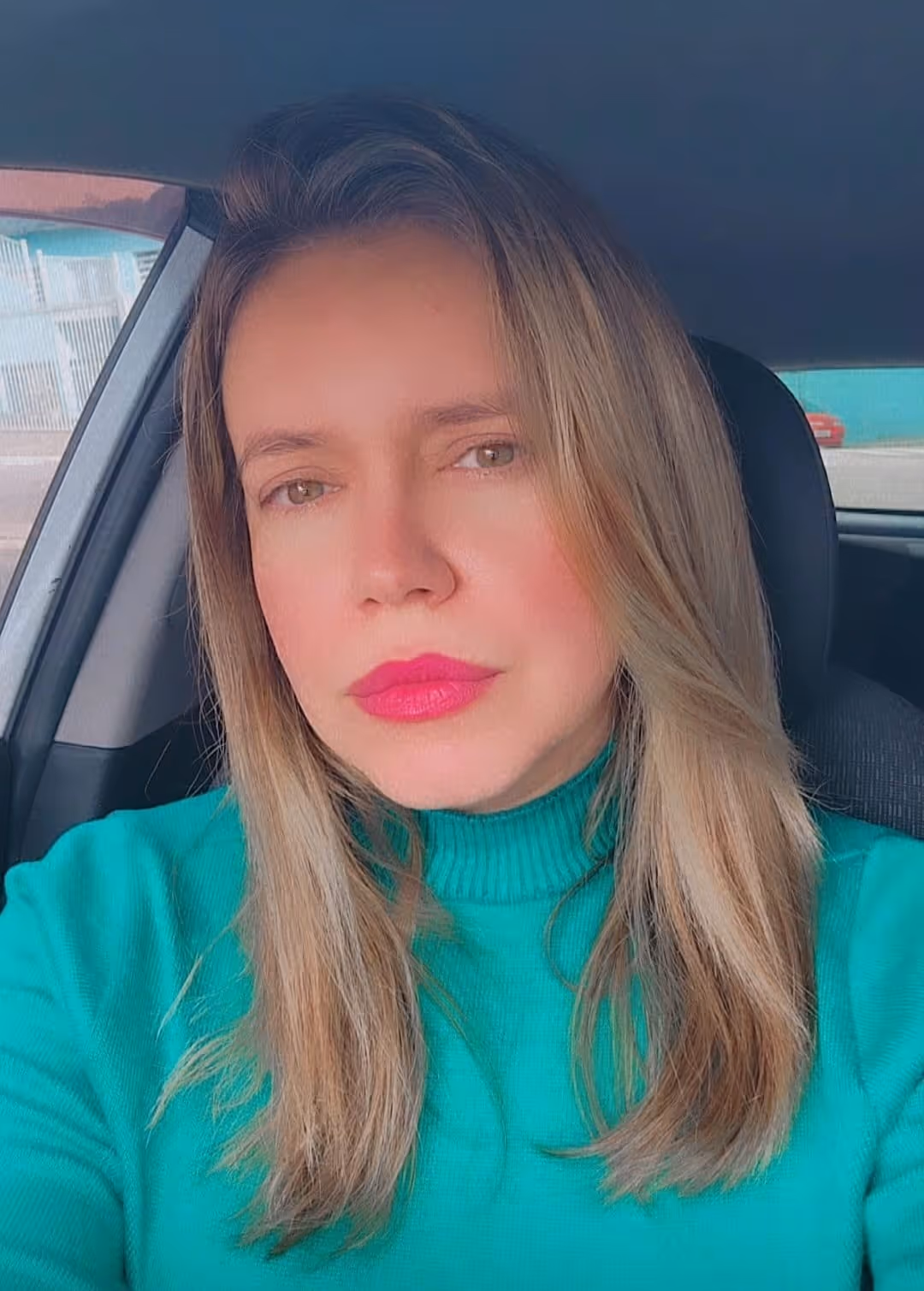 Mulher com cabelo loiro e batom rosa vibrante usando suéter verde sentado dentro de um carro.