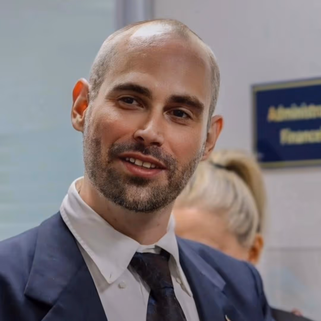 Homem careca com barba curta usando terno azul, gravata escura e camisa branca, sorrindo em ambiente interno.