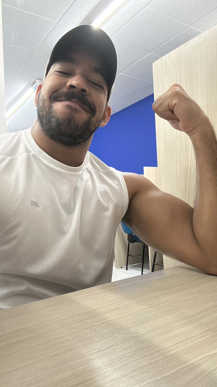 Homem sorridente usando boné preto e camiseta regata branca, flexionando o braço direito mostrando o bíceps.