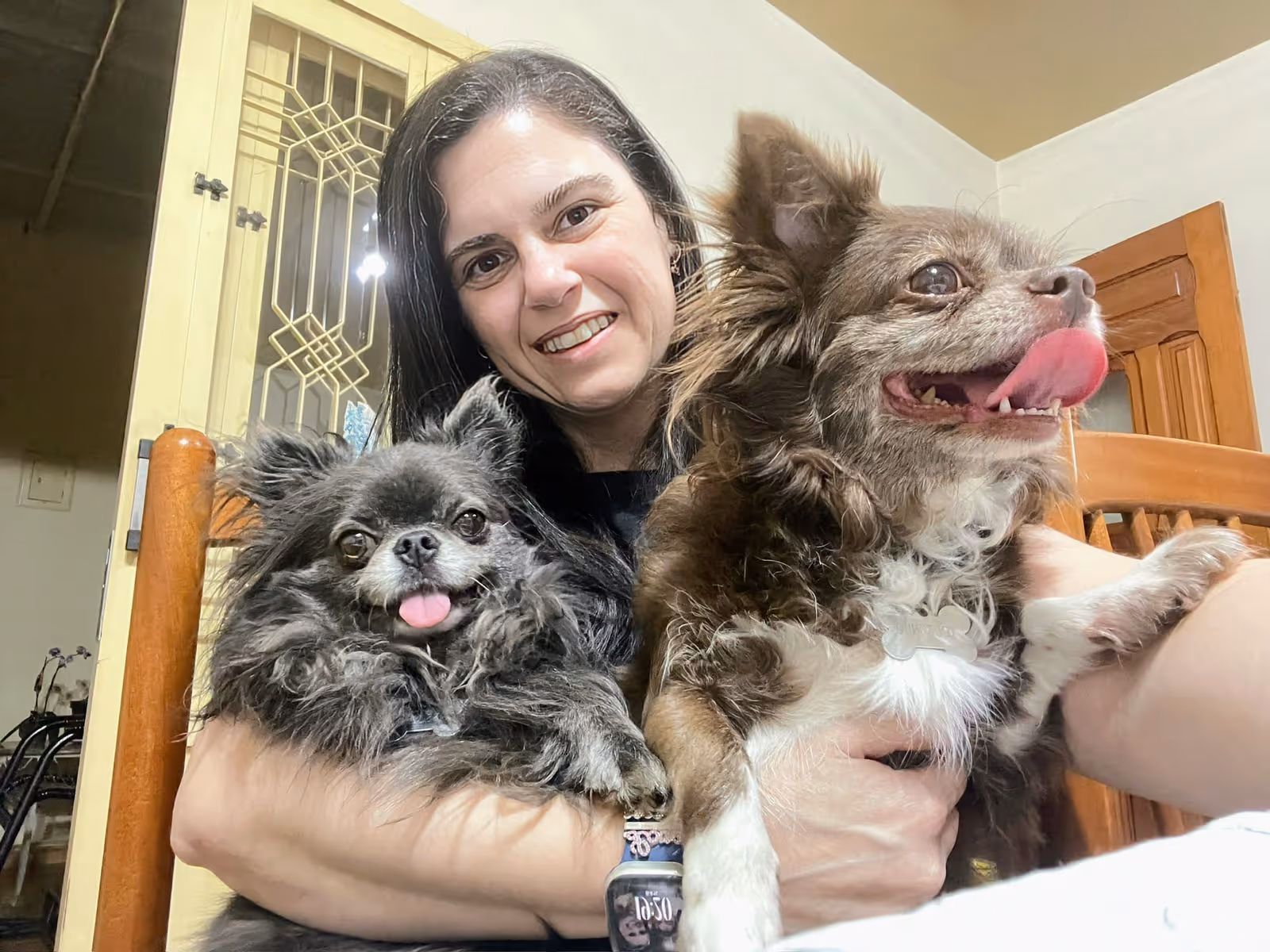 Mulher sorridente segurando dois cachorros pequenos, um com língua de fora e o outro lambendo o nariz.
