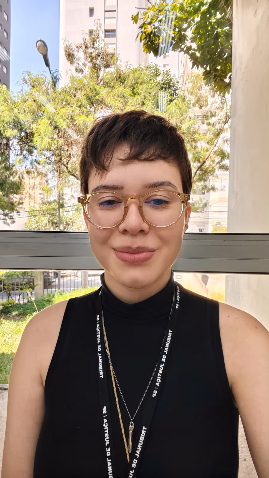 Pessoa com cabelo curto e óculos de armação clara, usando blusa preta sem mangas e colar com pingente, sorrindo levemente em ambiente urbano com árvores ao fundo.