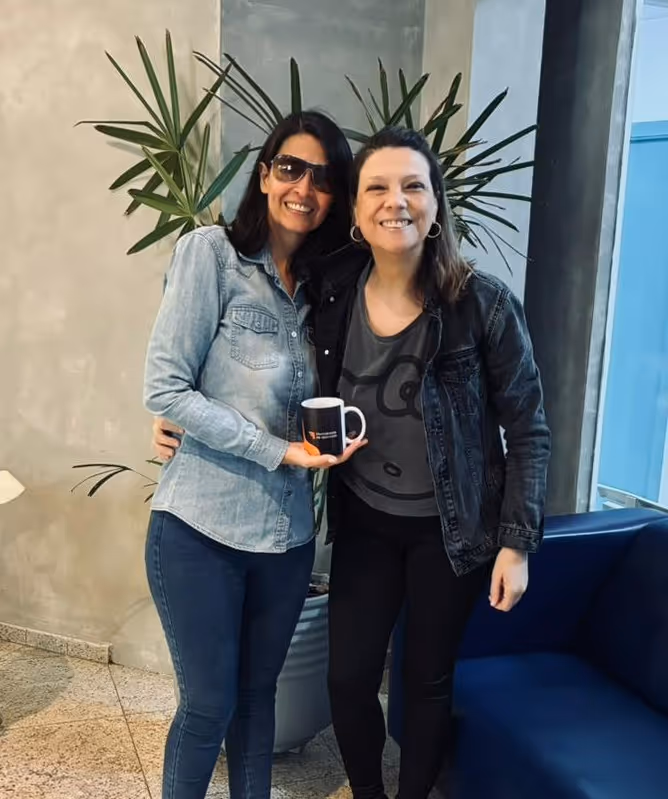Duas mulheres sorrindo e abraçadas em pé, uma segurando uma caneca preta, ao lado de uma planta alta e um sofá azul.