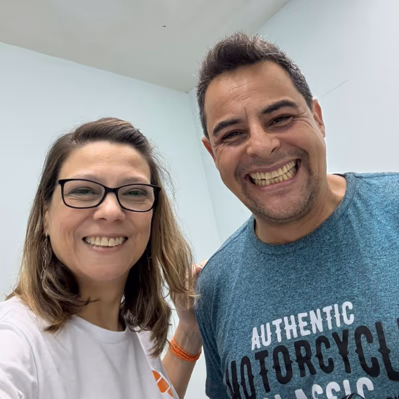 Duas pessoas sorrindo para a câmera, uma mulher de óculos e um homem com camiseta azul com texto sobre motocicletas.