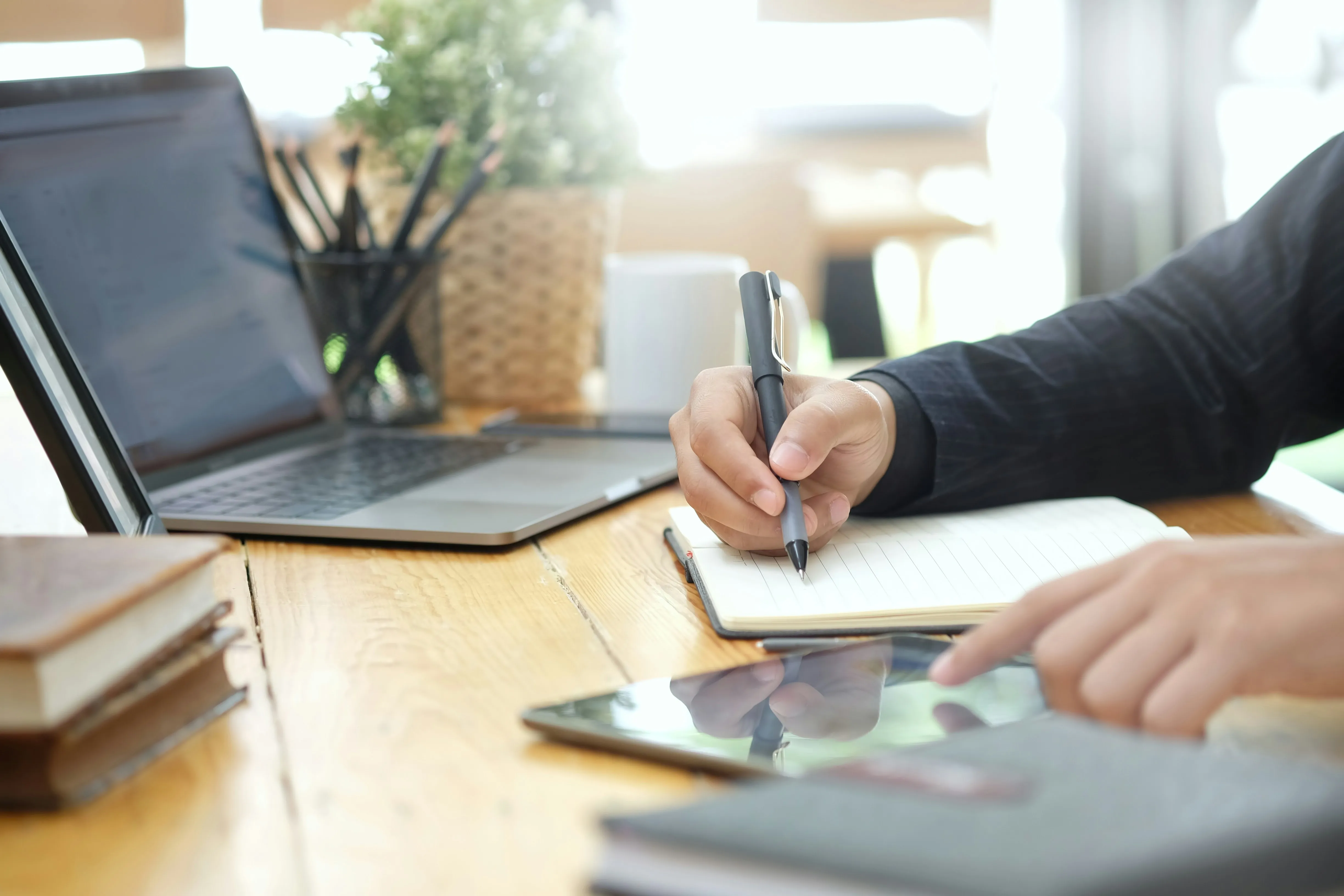Person writing in notebook at desk with laptop and books