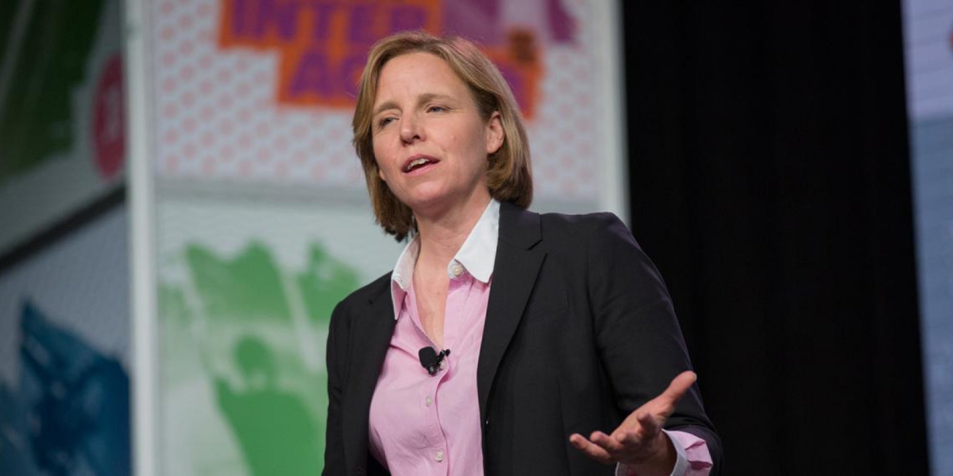 Megan Smith delivering a keynote at a tech conference, wearing a blazer and pink shirt, speaking passionately on stage.