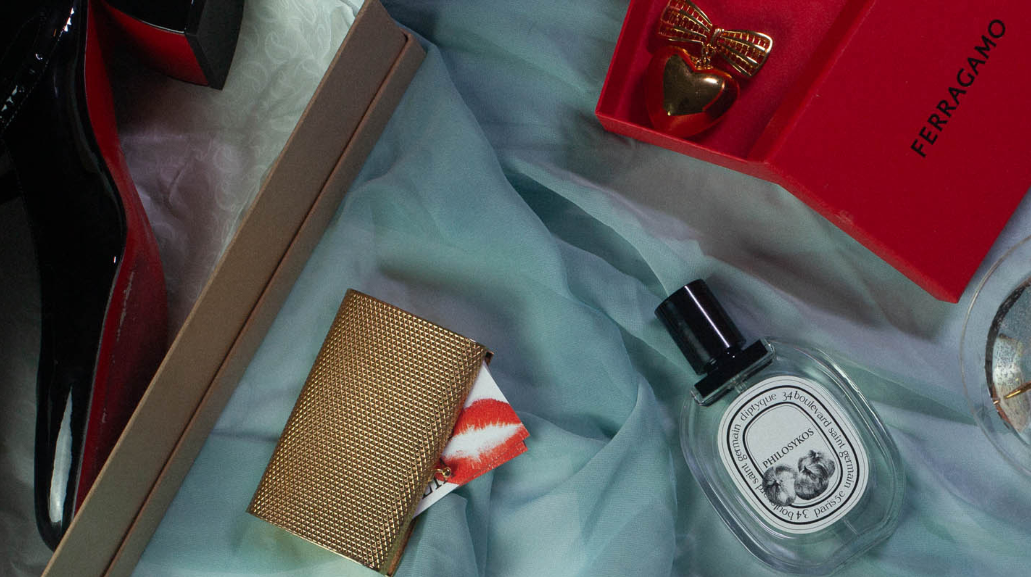 Close-up of Ferragamo gift box, Philosykos perfume, gold card holder, and black Louboutins on soft blue fabric with luxe textures.