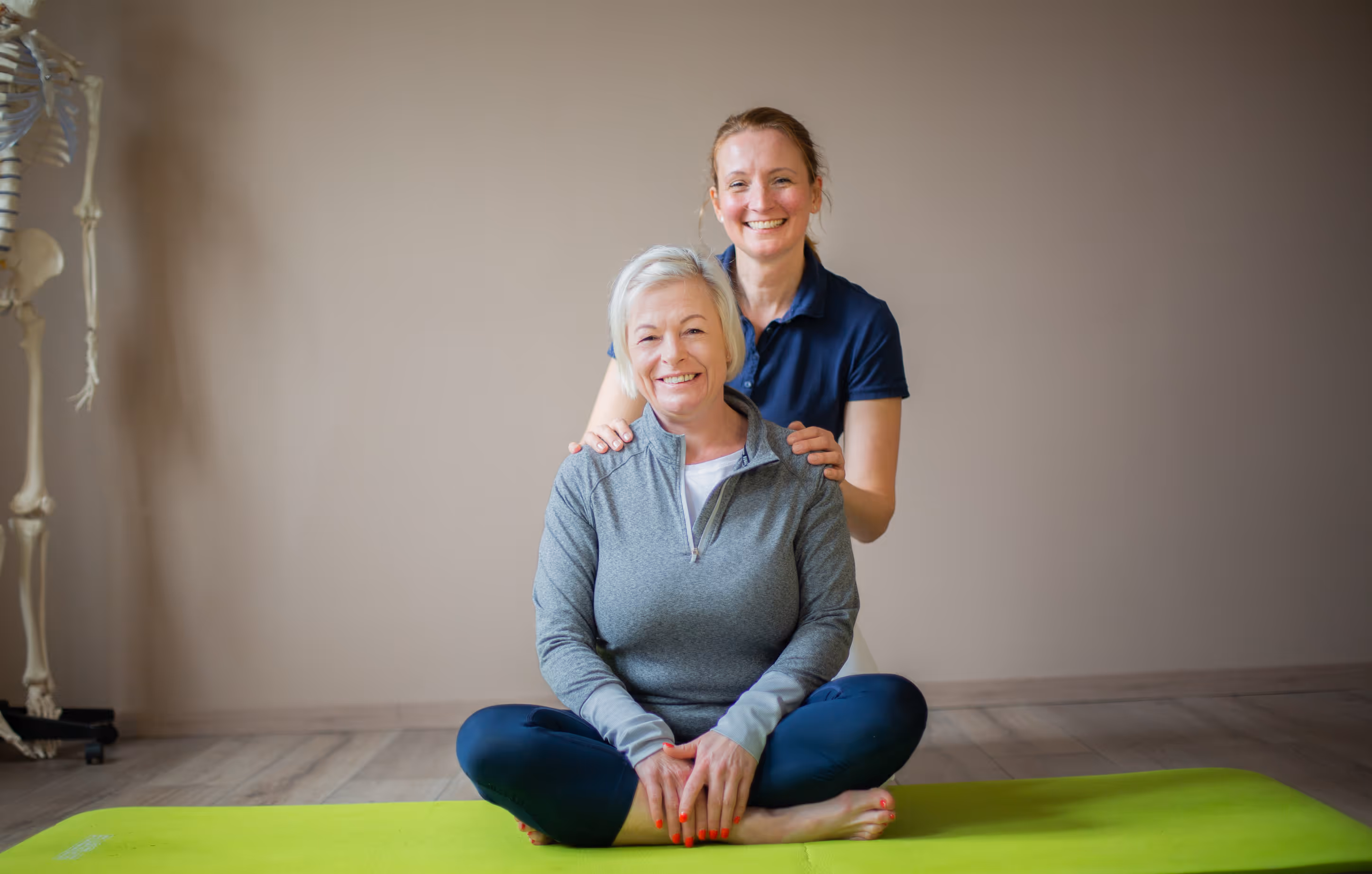 Zwei lächelnde Frauen, eine sitzt im Schneidersitz auf einer grünen Yogamatte, die andere steht hinter ihr und legt die Hände auf ihre Schultern.