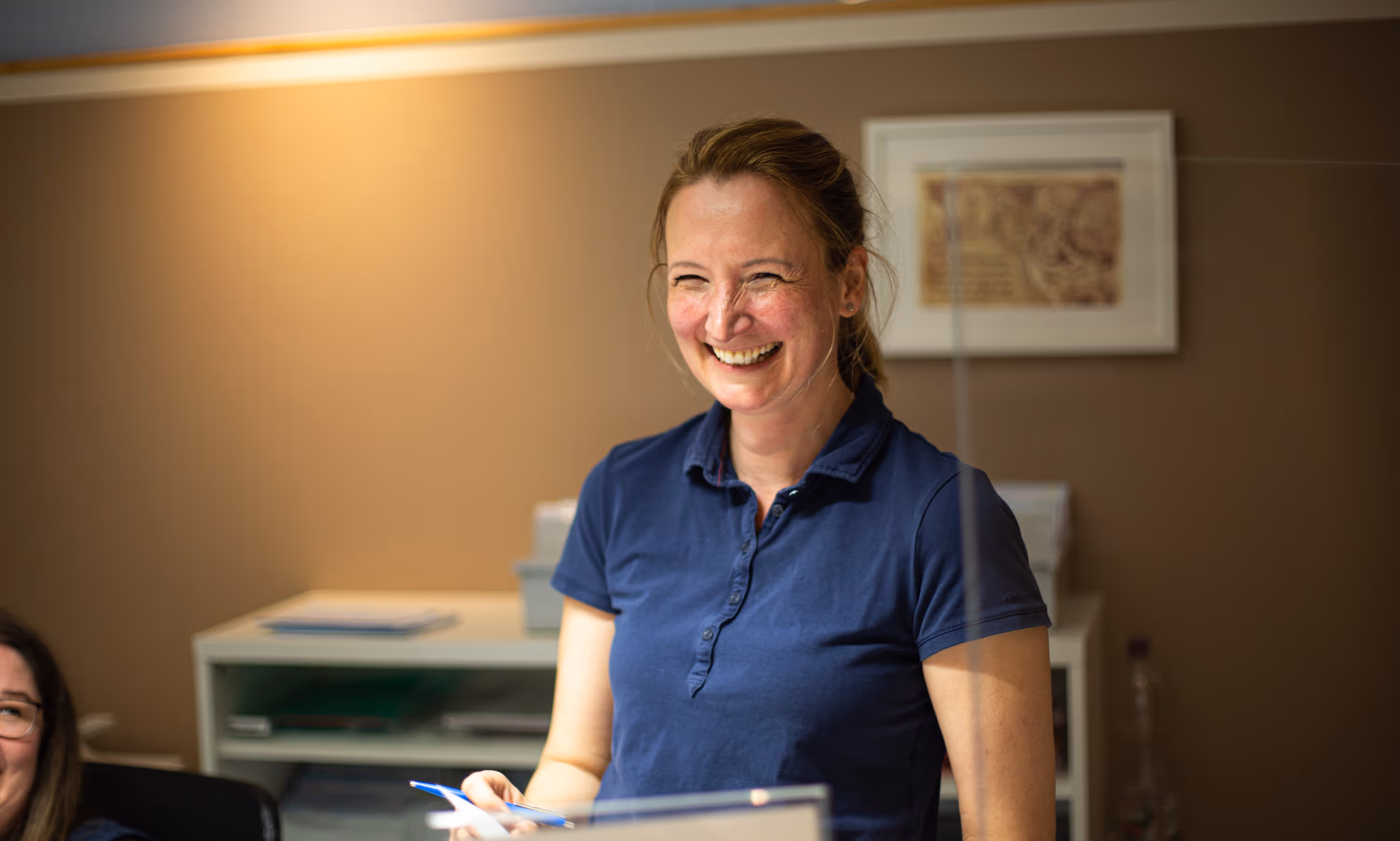 Lächelnde Frau in blauem Poloshirt steht in einem Büro mit einem Stift und Dokument in der Hand.
