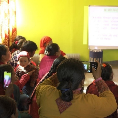 A group of people inside a bright room looking at a projector screen. A female farmer photographs a presentation on pest and disease management for future reference during the DFFS, while her friend attends with her infant
