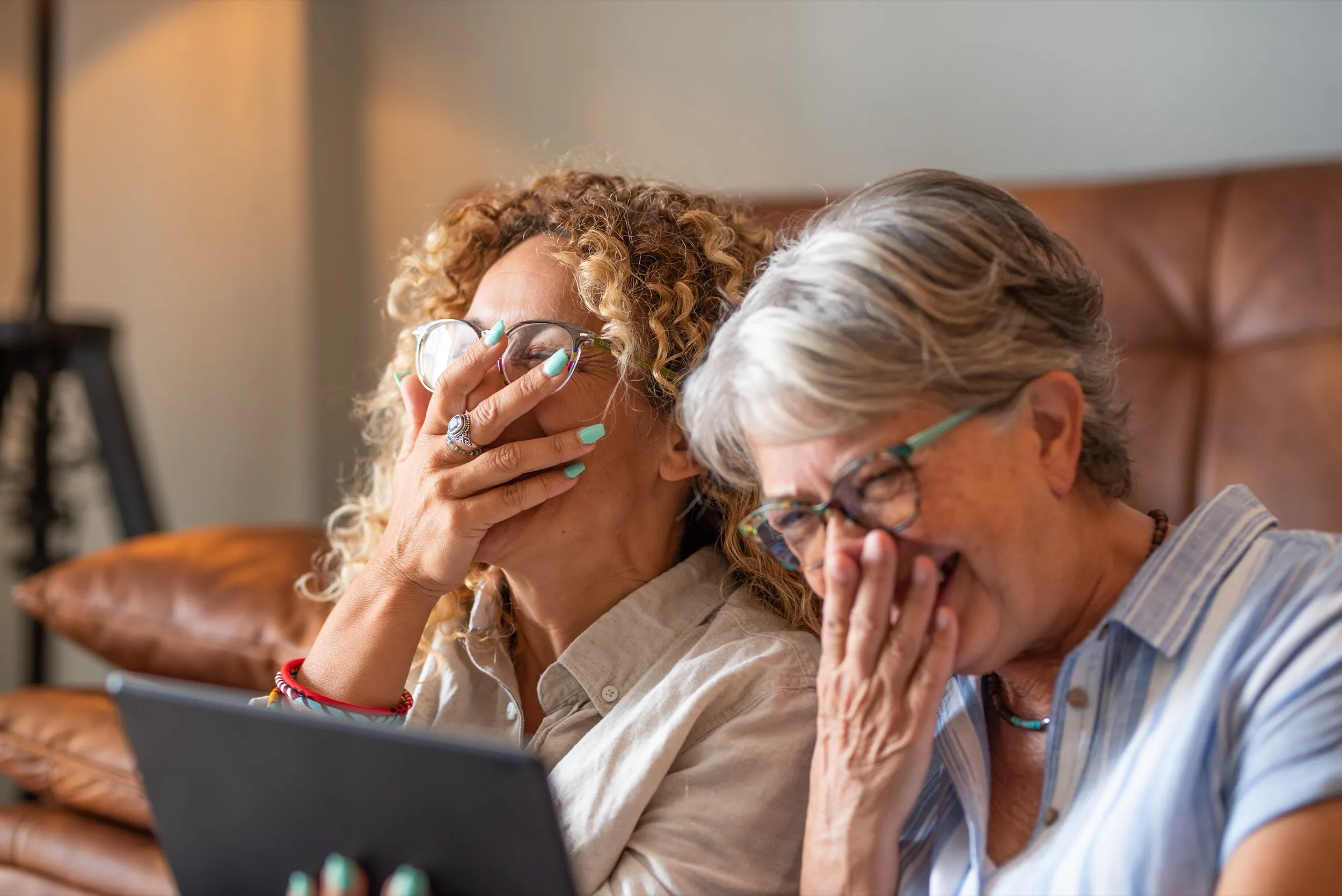 Two older women sitting on a leather couch laughing while looking at a tablet.
