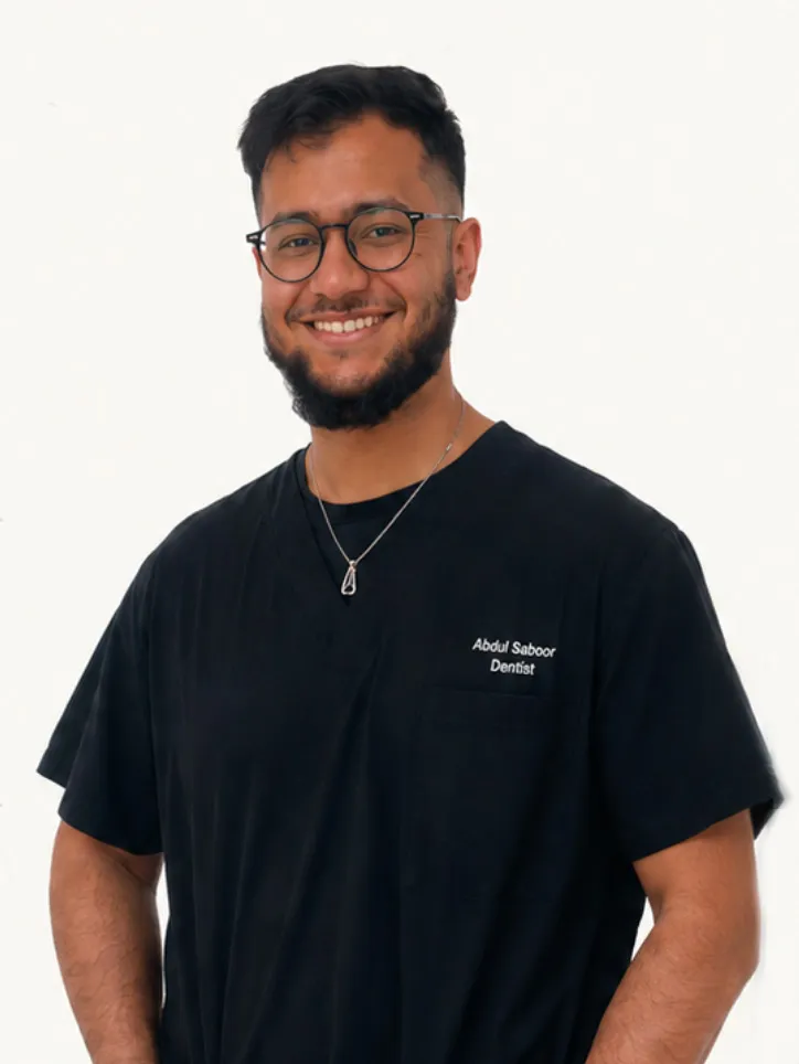 Smiling man with beard and glasses wearing a black dental scrub top embroidered with the name Abdul Saboor and the title Dentist, standing against a plain white background.