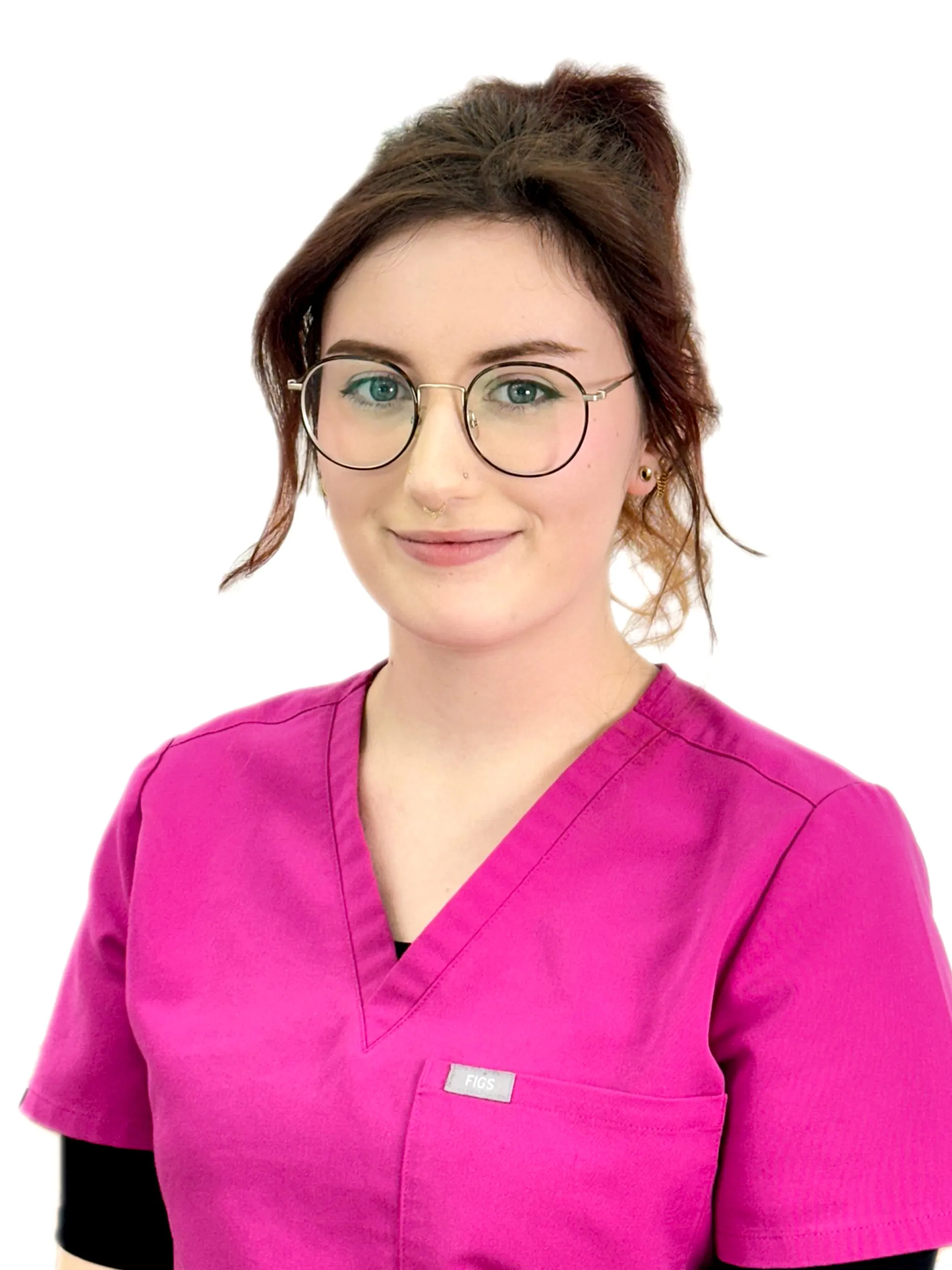 Young woman with light complexion, blue-green eyes, brunette hair in a loose bun, and round glasses wearing a bright pink scrub top over a black shirt.