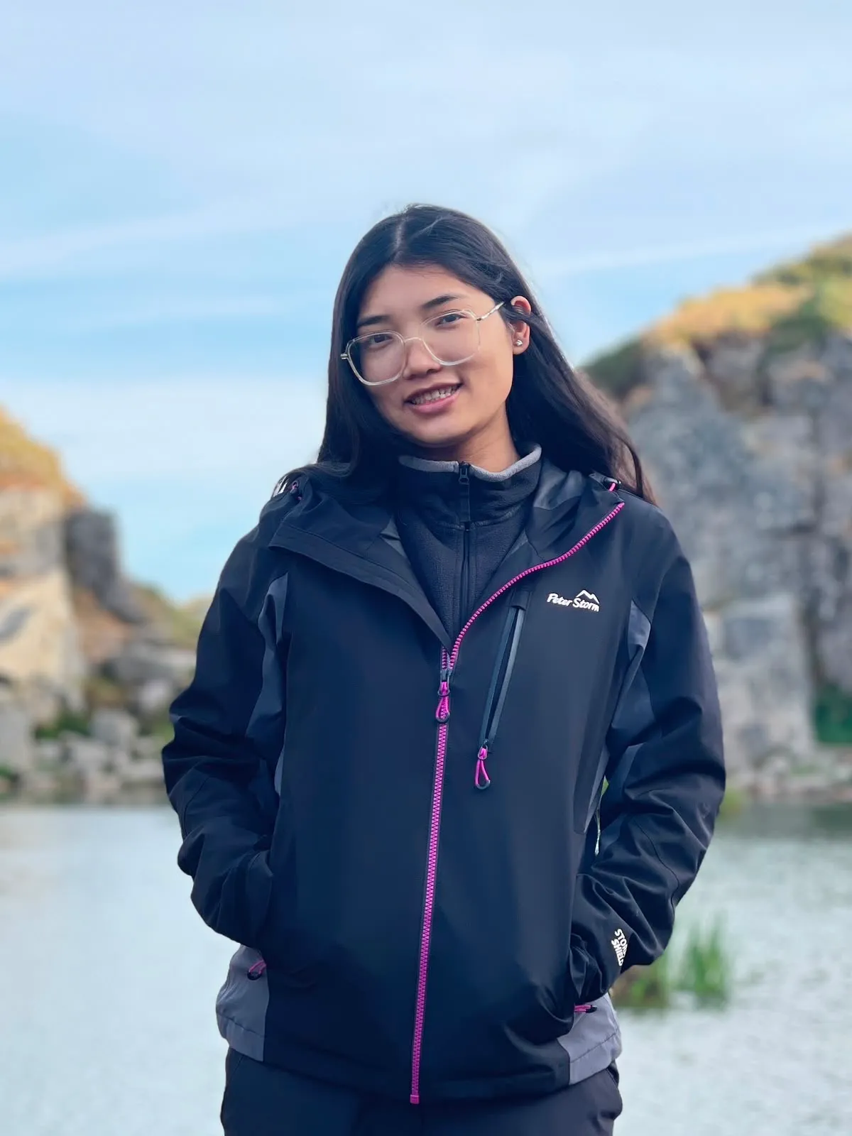 Young woman with long dark hair wearing clear glasses and a black jacket with purple zippers standing outdoors near a body of water and rocky terrain.